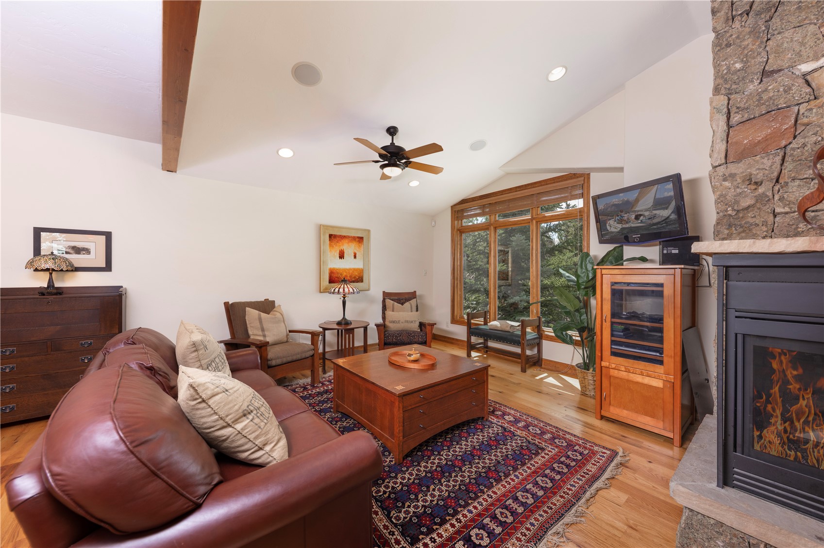 This cozy living room features a warm, rustic aesthetic with a leather sofa, a large wooden coffee table, and a patterned area rug. A stone-clad chimney breast anchors the space, housing a fireplace that adds to the inviting atmosphere. Large windows with wooden frames allow natural light to fill the room, highlighting the light wood flooring and the vaulted ceiling with a central fan.