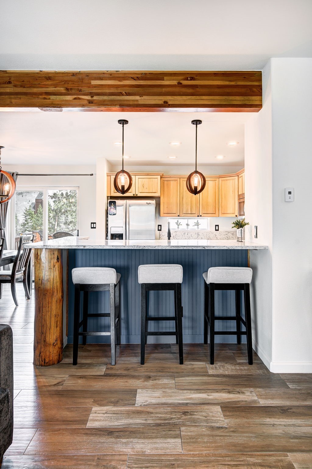 This kitchen features a central island with a blue paneled base and a light-colored stone countertop, complemented by three bar stools with dark frames and light upholstery. Above the island, two spherical pendant lights hang from the ceiling, while a rustic wooden beam adds a natural, textured element to the space. The kitchen includes light wood cabinetry, stainless steel appliances, and a view through a sliding glass door that leads to an outdoor area.