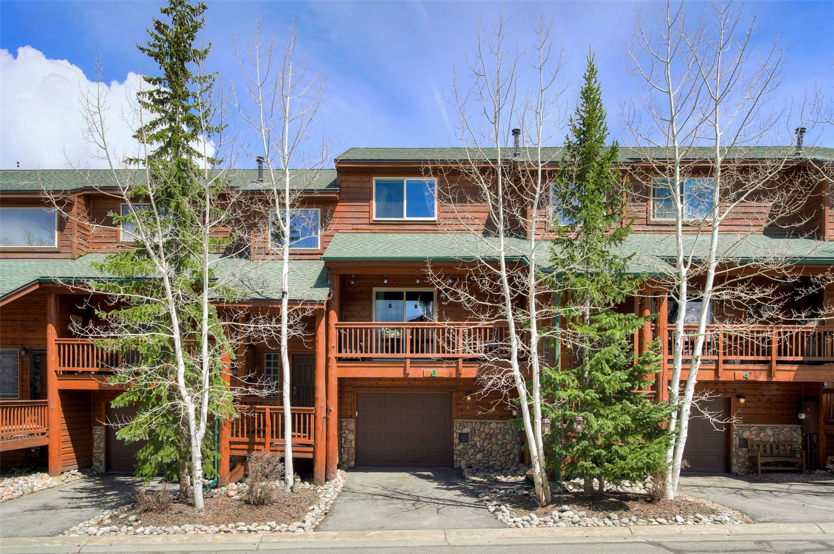 This image captures the front facade of a multi-unit mountain-style townhouse complex, featuring warm wood siding and green shingled roofs. Each unit includes a private garage on the ground level, a second-story balcony with wooden railings, and stone accents around the base. The scene is framed by mature aspen trees and evergreens, creating a rustic and inviting atmosphere under a clear blue sky.