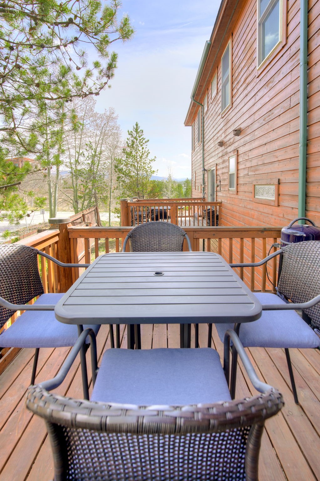 This image captures a spacious wooden deck featuring a central dining set with a dark metal table and four woven chairs with blue cushions. The perspective looks out from the deck toward a scenic, tree-lined landscape under a bright, clear sky. The side of the wooden house is visible on the right, showcasing its rustic siding and multiple windows, creating an inviting outdoor living space.