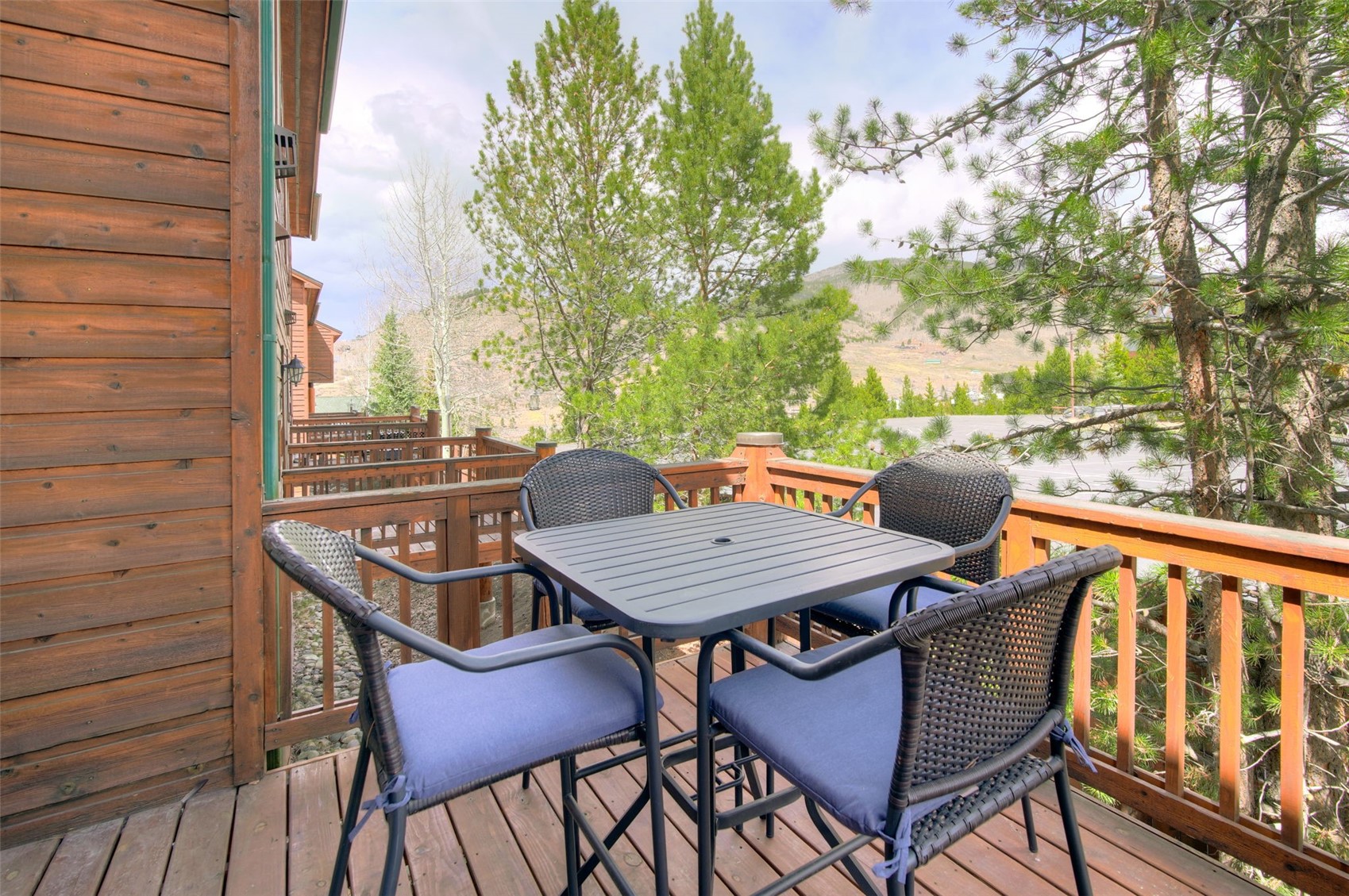 This elevated wooden deck features a casual dining set with four wicker-style chairs and a square metal table, perfect for outdoor relaxation. The space is enclosed by a sturdy wooden railing and offers a scenic view of surrounding pine trees and a distant mountain landscape. The warm wood tones of the deck and siding create a cozy, rustic atmosphere typical of a mountain retreat.