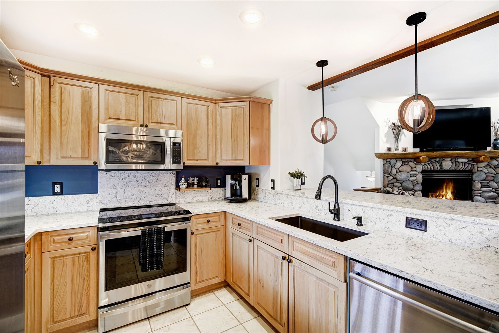 This kitchen features light wood cabinetry paired with light-colored stone countertops and a dark blue backsplash. The space is equipped with stainless steel appliances, including a microwave and range, and includes a dark sink with a modern faucet. A view into the adjacent living area reveals a stone-clad fireplace, creating an open and connected floor plan.