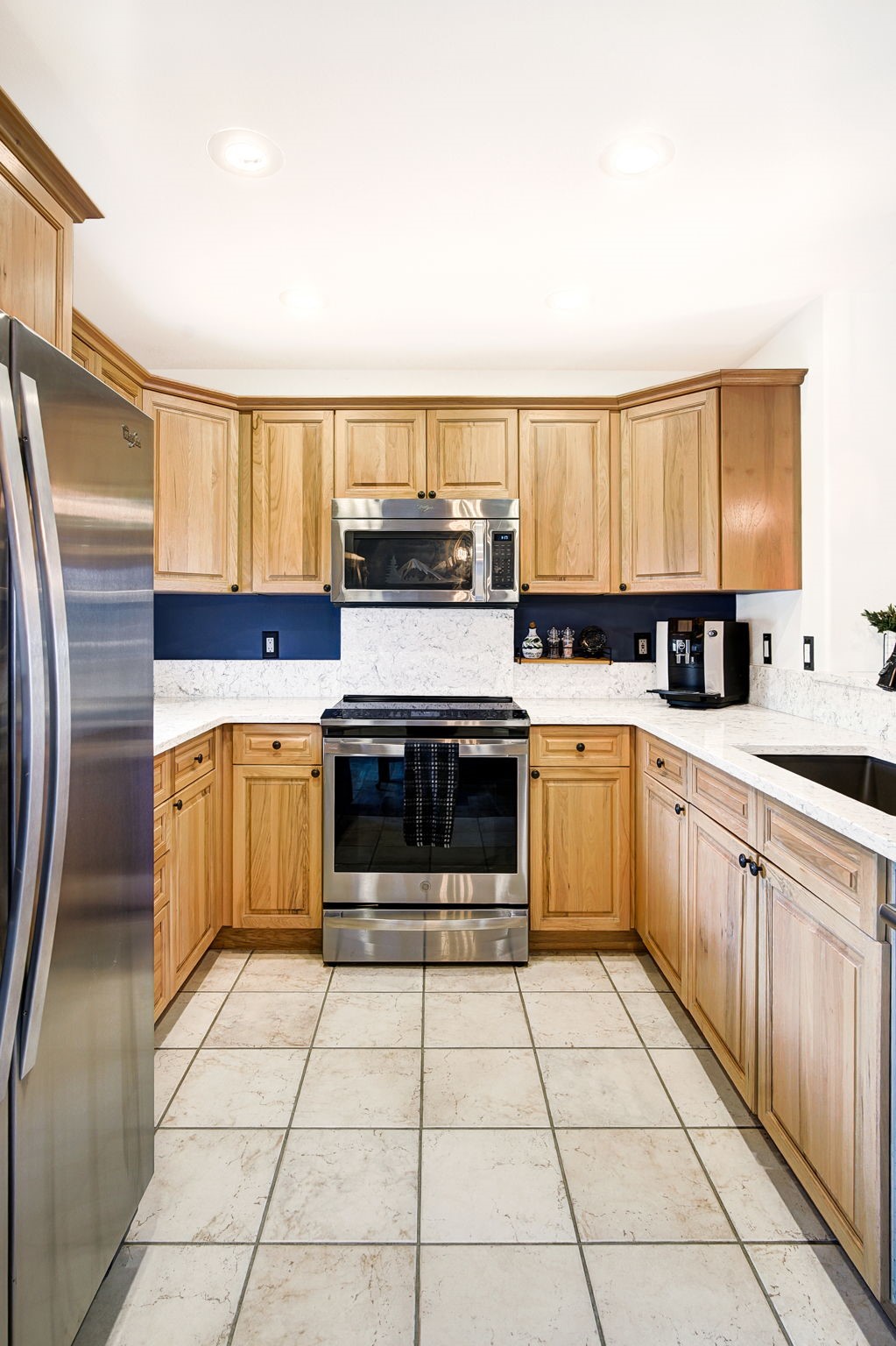 This U-shaped kitchen features light wood cabinetry paired with a bold dark blue backsplash and light-colored stone countertops. The space is equipped with stainless steel appliances, including a built-in microwave and a range, and is finished with neutral-toned square floor tiles. The perspective is a centered, eye-level shot looking directly into the workspace, highlighting the functional layout and clean aesthetic.