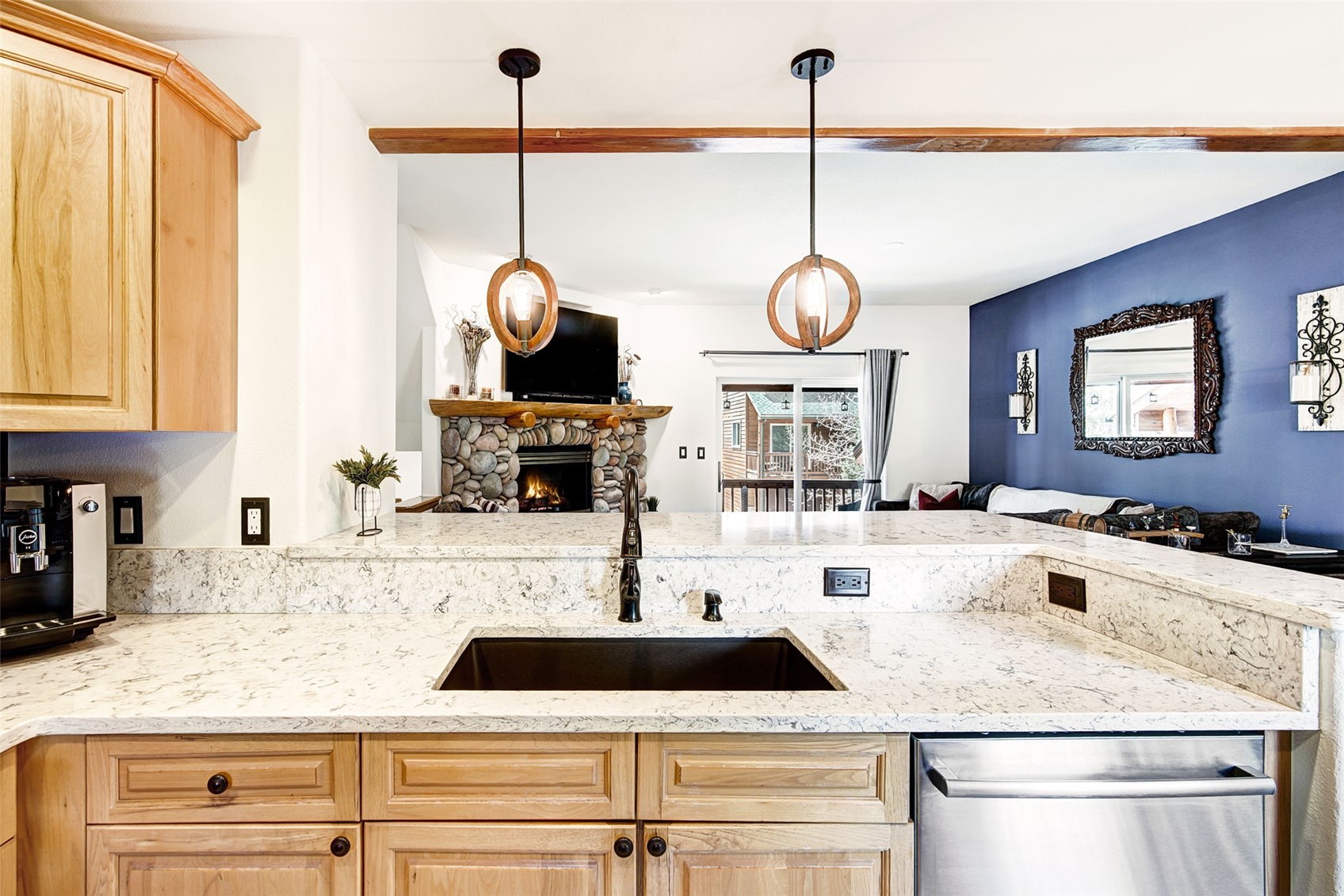 This kitchen features light wood cabinetry paired with light-toned stone countertops and a dark, rectangular undermount sink. Two spherical pendant lights hang above the breakfast bar, which overlooks an open-concept living area with a stone-clad fireplace and a deep blue accent wall. The perspective is from the kitchen workspace, looking out toward the living room and a sliding glass door that leads to an outdoor deck.