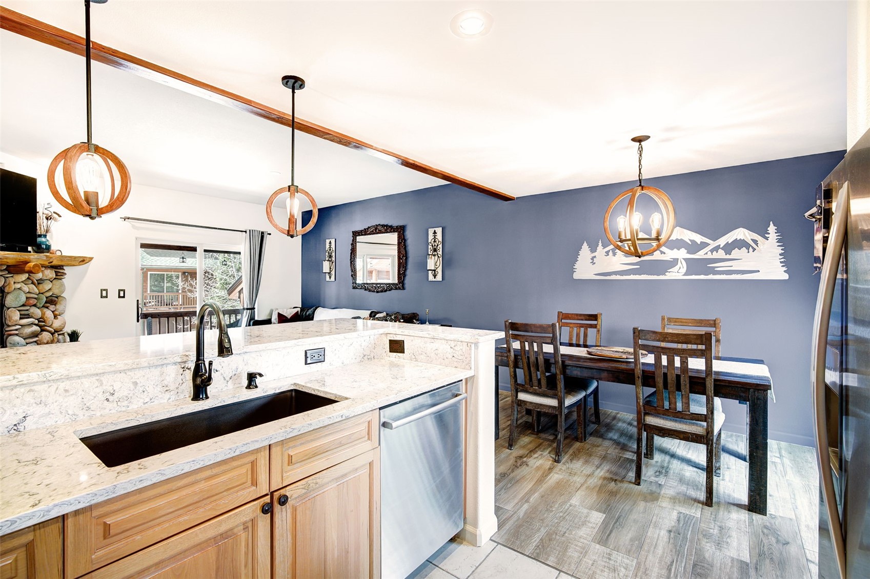 This open-concept kitchen features light wood cabinetry paired with light-toned stone countertops and a dark undermount sink. A dining area sits adjacent to the kitchen, defined by a deep blue accent wall decorated with a mountain-themed wall art piece and illuminated by a spherical pendant light. The space is connected to a living area with a stone-clad chimney breast, creating a cohesive and inviting atmosphere.
