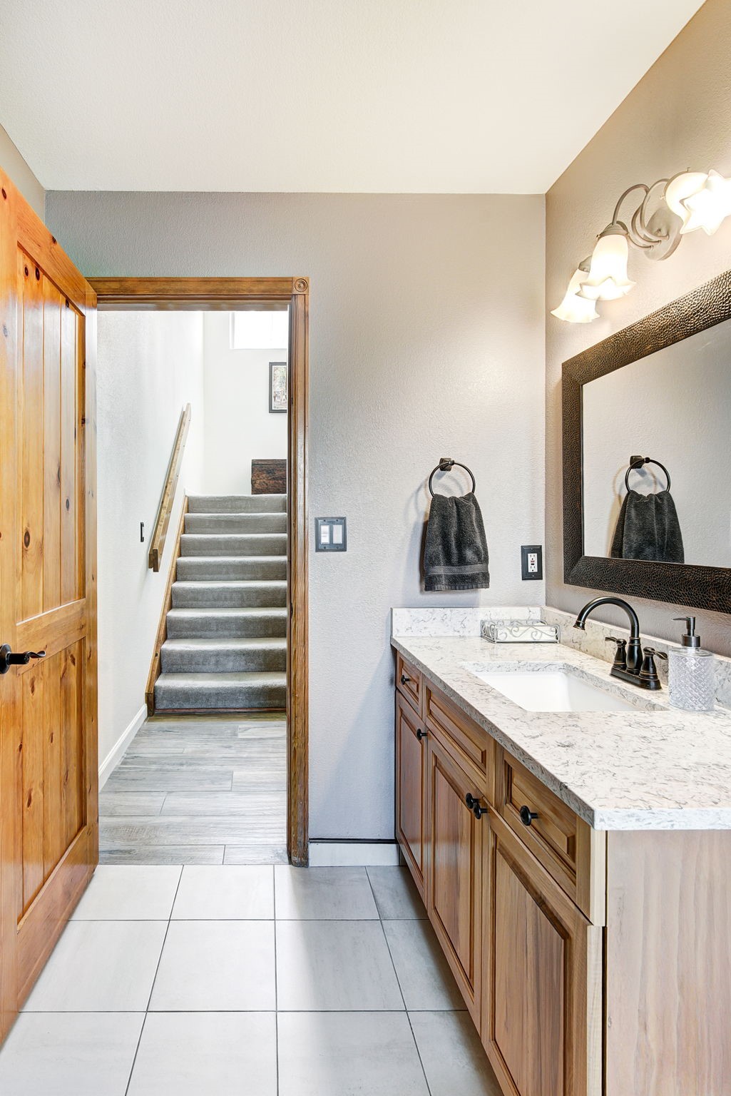 This bathroom features a vanity with a light-toned wood cabinet and a patterned stone countertop, complemented by a dark-framed mirror and matching hardware. The space includes a towel ring with a dark towel and a three-light vanity fixture above the mirror. A doorway leads directly to a staircase with carpeted steps, creating a transition between the tiled bathroom floor and the hallway area.