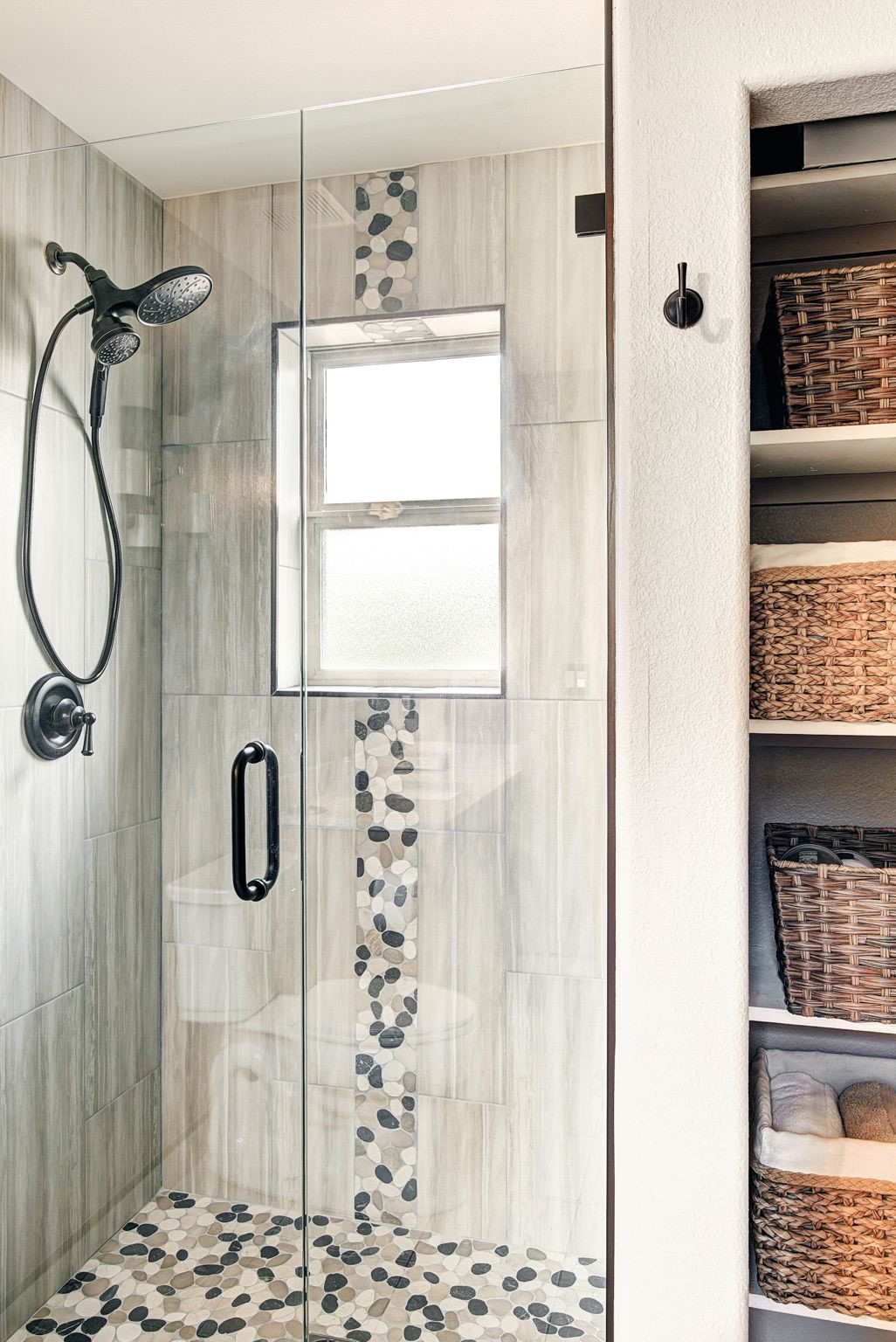 This bathroom features a walk-in shower enclosed by a glass door, showcasing a pebble-stone floor and a matching vertical accent strip on the wall. To the right, a built-in shelving unit provides organized storage with woven baskets, while a black wall hook adds a functional touch. The neutral color palette and clean lines create a modern and spa-like atmosphere.