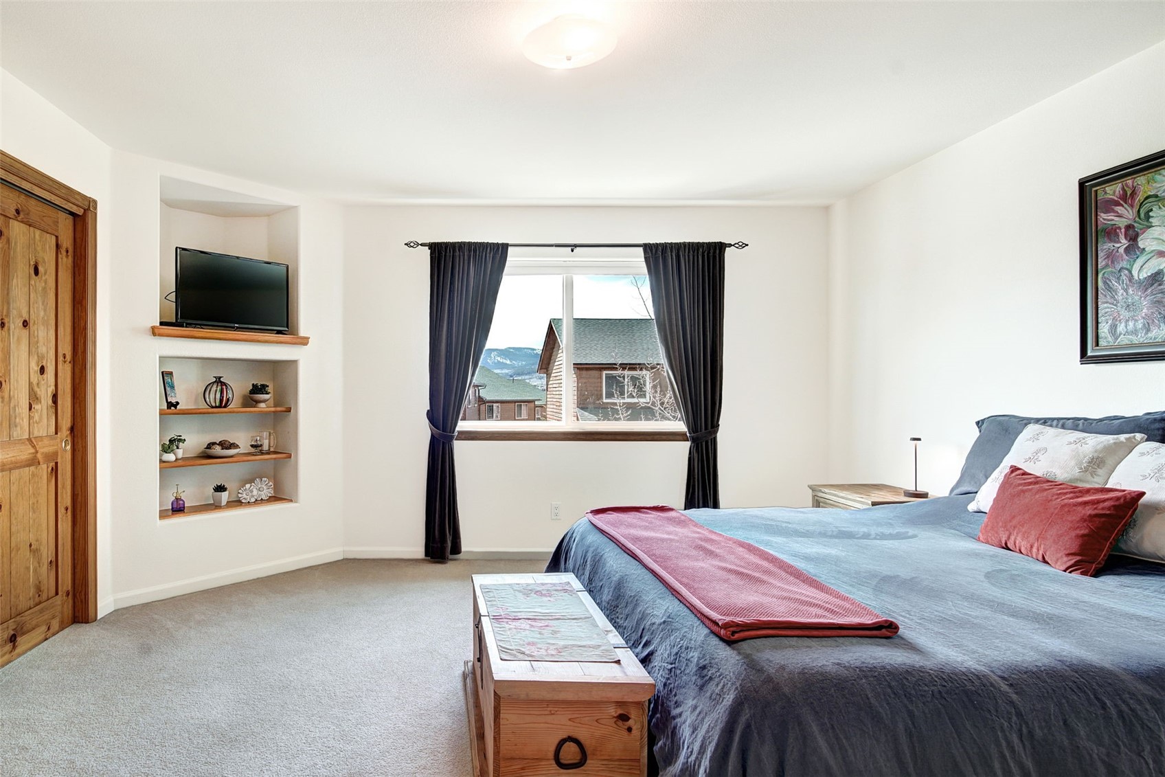 This primary bedroom features a neutral color palette with a large bed dressed in dark blue bedding and a red throw blanket. To the left, a built-in shelving unit provides storage and display space, topped with a television, while a wooden chest sits at the foot of the bed. A central window framed by dark curtains offers a view of the surrounding landscape, and a framed piece of art hangs on the wall above the bed.