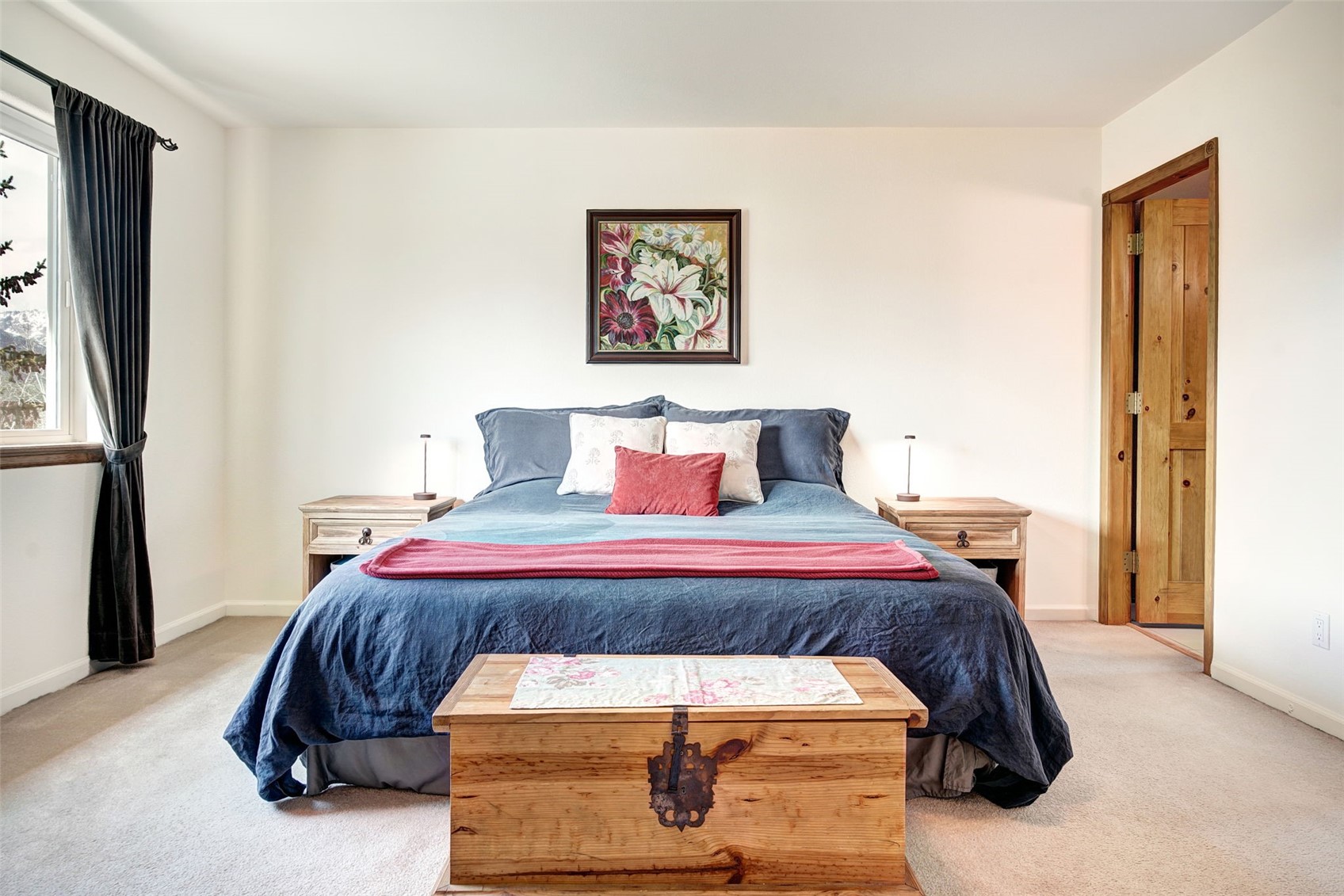 This primary bedroom features a centered bed dressed in a deep blue duvet with a matching red throw blanket and decorative pillows. A rustic wooden chest sits at the foot of the bed, while matching light wood nightstands with minimalist lamps flank the headboard. A floral-themed painting hangs above the bed, and a wooden door leads to an adjacent space, creating a warm and inviting atmosphere.