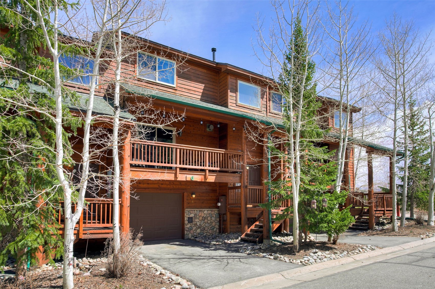 This multi-story townhome features a rustic wood-sided exterior with a prominent second-story balcony and a ground-level garage. The facade is accented by stone masonry around the garage entrance and is framed by mature aspen trees under a clear blue sky. The perspective is a street-level view capturing the inviting mountain-style architecture and the paved driveway leading to the home.