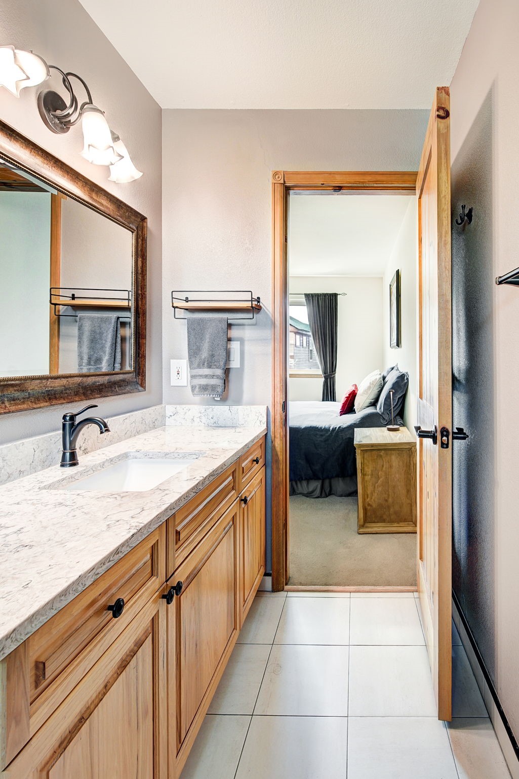 This bathroom features a light wood vanity topped with a patterned stone countertop and a dark metal faucet. A large mirror with a decorative frame hangs above the vanity, reflecting the space, while a doorway leads directly into a bedroom with a neatly made bed. The room is finished with light-colored floor tiles and neutral wall paint, creating a clean and functional transition between the two living areas.