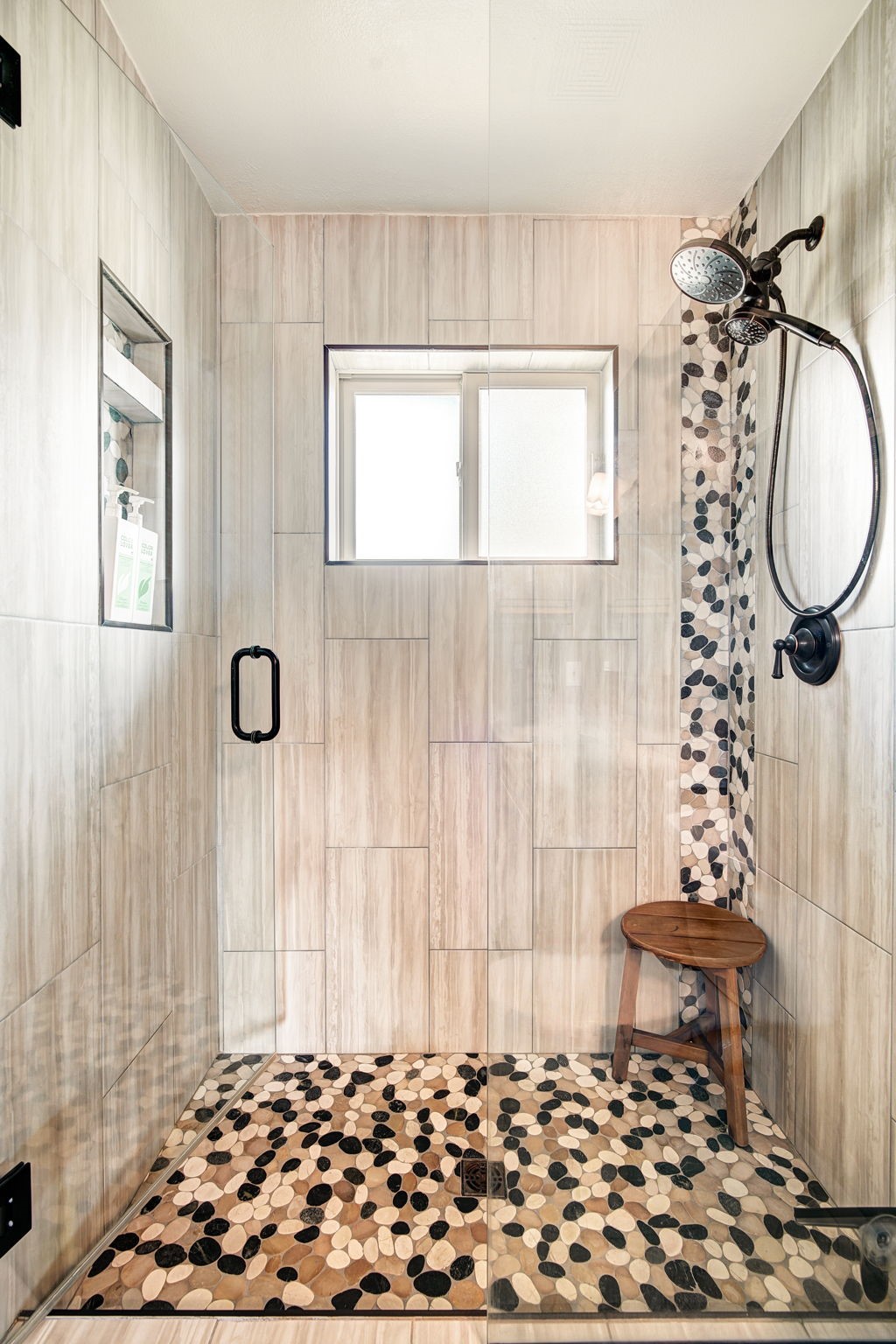 This walk-in shower features a contemporary design with light-toned, vertical-grain wall tiles and a striking pebble-stone floor. A glass enclosure provides a clear view of the interior, which includes a built-in recessed niche for storage, a small wooden stool, and a dark-finished shower fixture. The space is illuminated by a central window, creating a bright and spa-like atmosphere.
