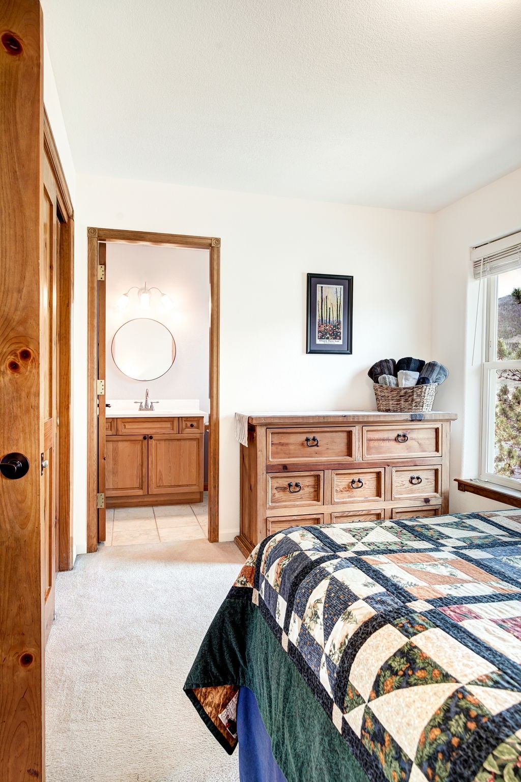 This bedroom features a cozy, traditional aesthetic with a patchwork quilt on the bed and a light wood dresser topped with a woven basket. A doorway leads directly into an adjoining bathroom, which is visible and includes a vanity with a round mirror and light fixture. The room is brightened by natural light from a window on the right, creating a warm and inviting atmosphere.