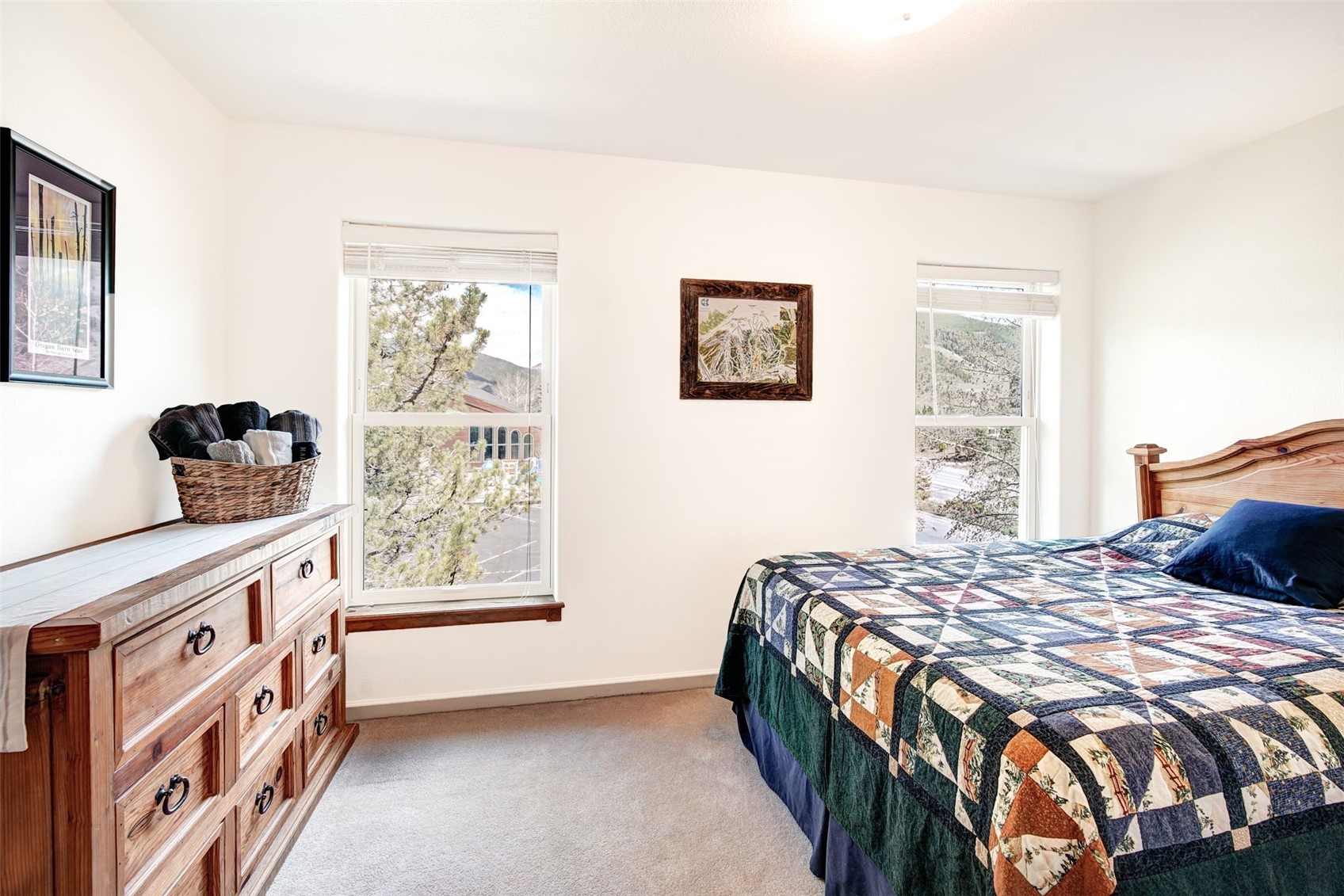 This cozy bedroom features a wooden bed frame dressed in a patterned quilt and a matching wooden dresser topped with a woven basket. Two windows provide natural light and views of the surrounding landscape, while framed artwork adds a personal touch to the neutral walls. The room is carpeted and offers a comfortable, rustic atmosphere.