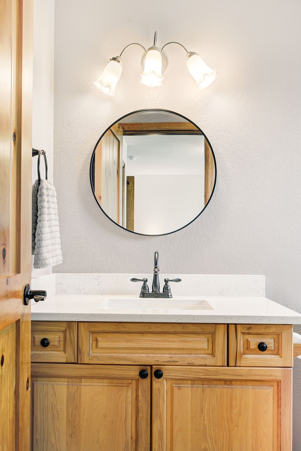 This bathroom features a light wood vanity topped with a light-colored stone countertop and a centered sink. A round mirror with a dark frame hangs above the vanity, centered beneath a three-light fixture with frosted glass shades. The space is finished with neutral-toned walls and includes a towel hanging on the left, creating a clean and functional aesthetic.
