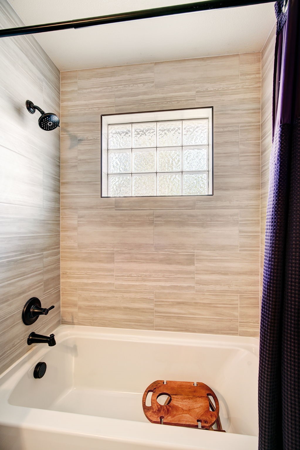This bathroom features a clean, white bathtub equipped with matte black fixtures, including a showerhead and faucet. The wall surrounding the tub is finished with horizontal, light-toned textured tiles and includes a glass block window that allows for natural light while maintaining privacy. A wooden bath tray rests inside the tub, adding a warm, spa-like touch to the neutral space.