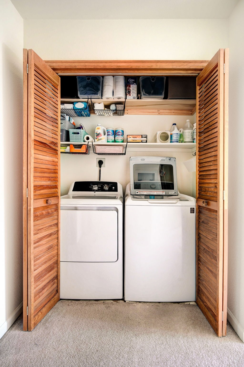 This laundry closet features a side-by-side washer and dryer set tucked behind classic louvered wood doors. Above the appliances, two levels of shelving provide organized storage with wire baskets and various household supplies. The space is compact and functional, utilizing vertical storage to maximize the utility of the closet area.