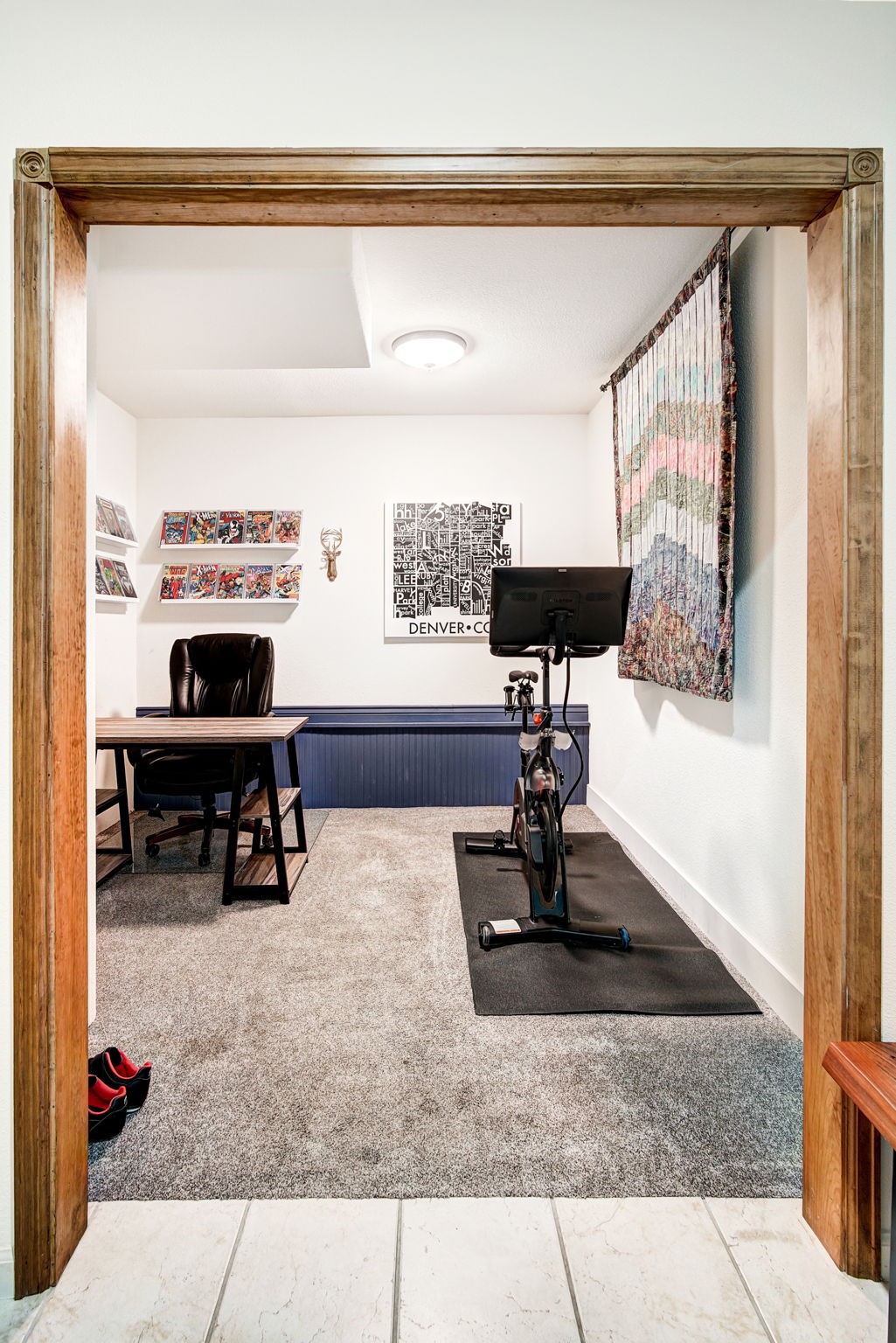 This multi-functional room serves as both a home office and a fitness space, viewed through a wide, wood-trimmed doorway. The area features gray carpeting, a desk with a black leather chair, and a stationary exercise bike positioned on a black rubber mat. Decorative elements include wall-mounted comic book displays and a framed Denver map print, creating a personalized and versatile living space.