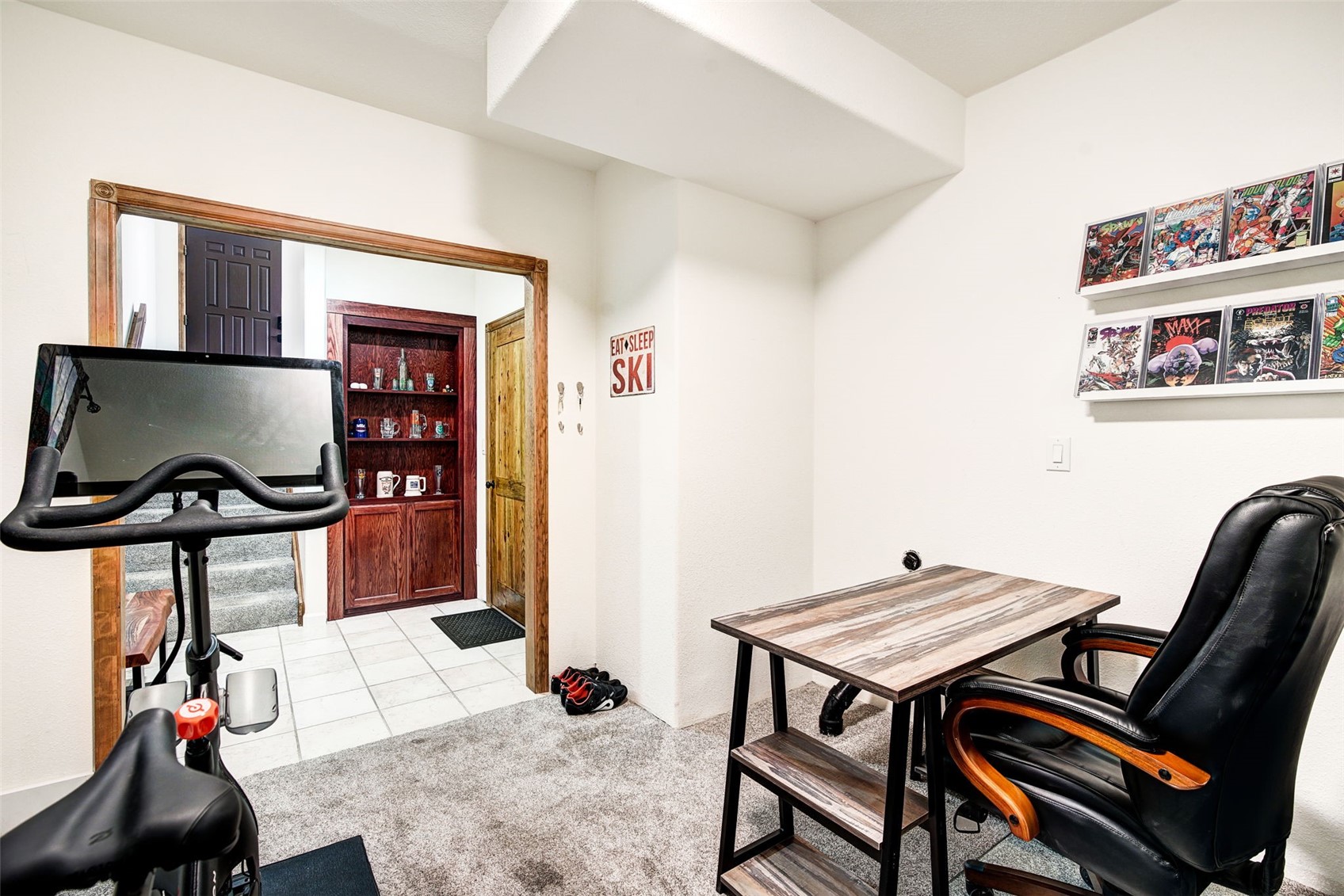 This multi-functional space serves as both a home gym and a compact office area, featuring a stationary exercise bike in the foreground and a small desk with a leather office chair. The room transitions into a tiled hallway that leads to a staircase and a built-in wooden shelving unit, while a wall-mounted display of comic books adds a personalized touch. The neutral walls and carpeted flooring create a versatile environment suitable for various activities.