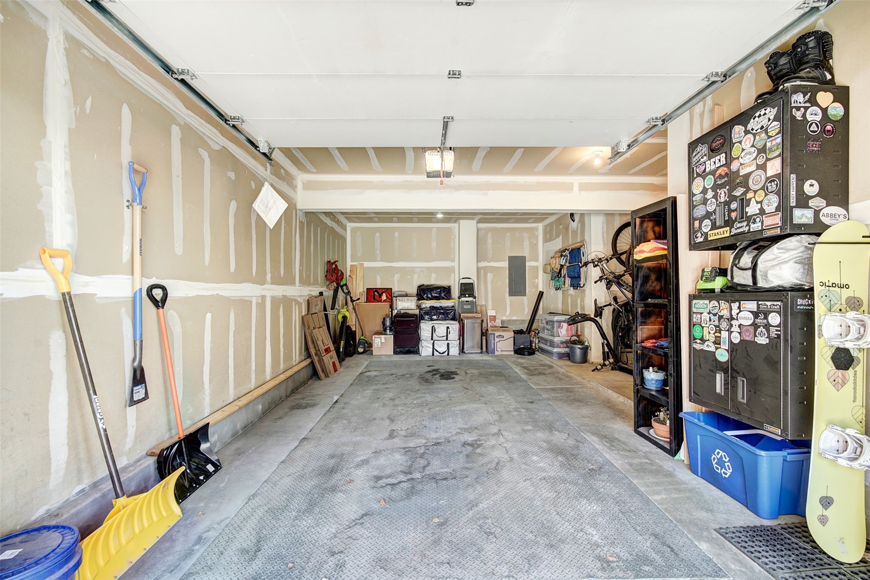 This image shows the interior of a spacious, unfinished garage with exposed drywall seams and a textured concrete floor. The space is utilized for storage, featuring a tall metal locker covered in stickers, a shelving unit, bicycles, and various tools leaning against the walls. The perspective is a wide-angle shot from the garage door looking inward, capturing the depth and functional layout of the storage area.