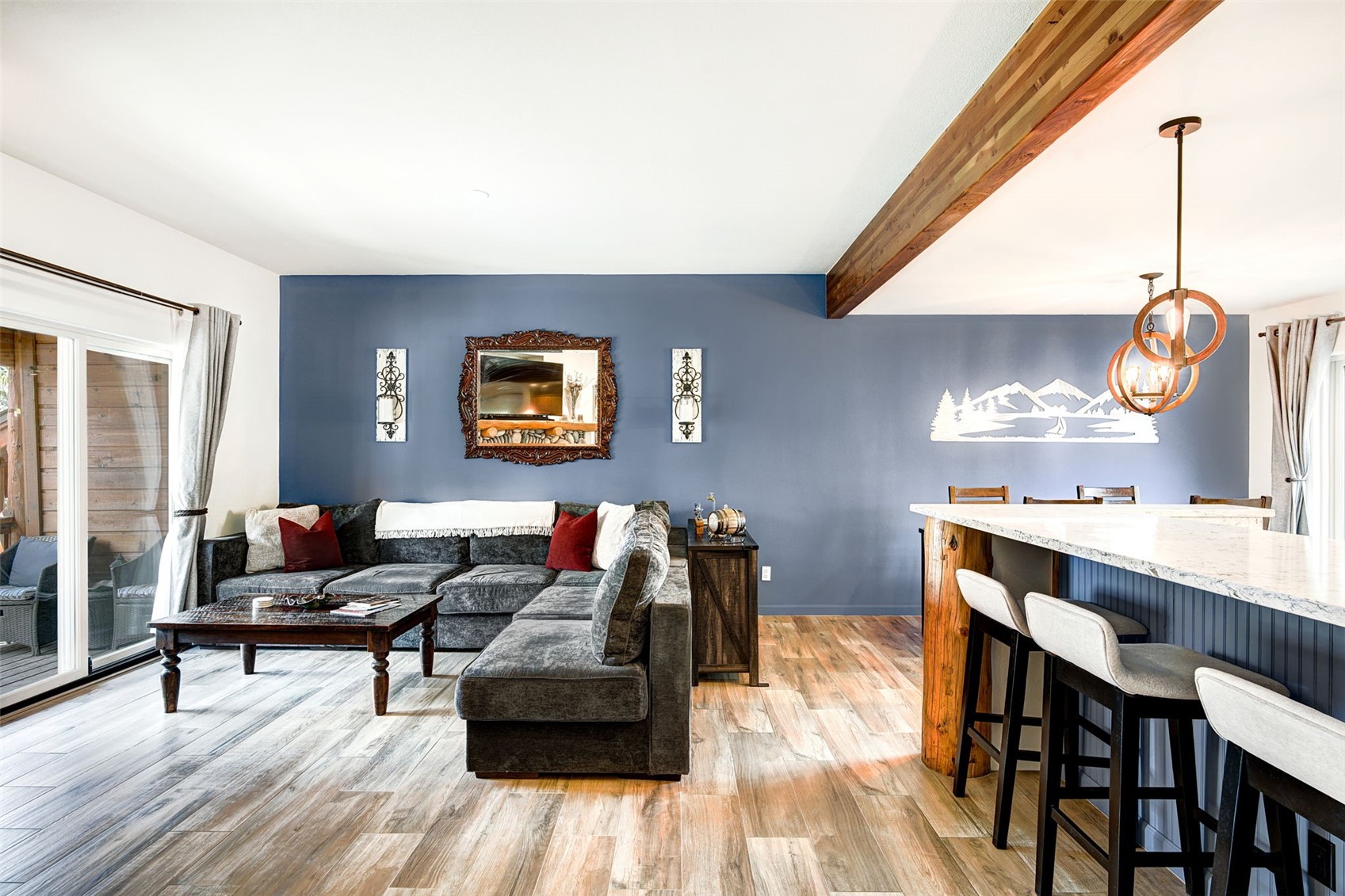 This open-concept living area features a deep blue accent wall adorned with a decorative mirror and wall sconces, creating a cozy focal point. A large, dark-toned sectional sofa sits atop wood-look flooring, complemented by a rustic coffee table and a nearby kitchen island with bar seating. The space is brightened by natural light from sliding glass doors and warm pendant lighting, offering a comfortable and inviting atmosphere.