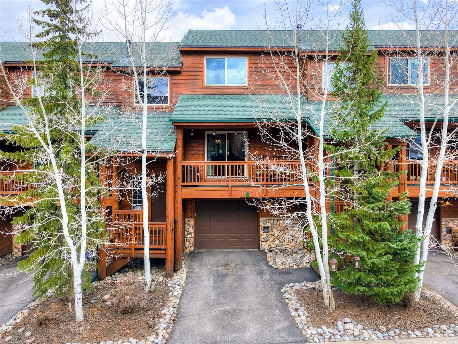 This mountain-style townhome features a rustic wood-paneled facade with a green shingled roof and a stone-accented lower level. The property includes a private garage, a wooden balcony deck, and a small front porch area, all framed by mature aspen and evergreen trees. The perspective is a straight-on eye-level shot, capturing the inviting, cozy aesthetic of a woodland retreat.