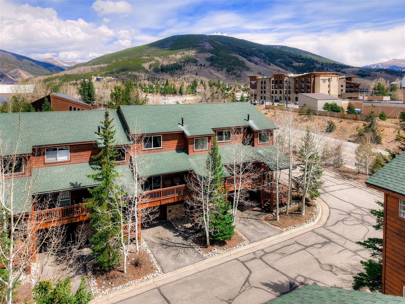 This elevated perspective captures a multi-level mountain townhome complex featuring classic wood siding and green shingled roofs. The property includes private balconies, covered parking areas, and mature aspen and pine trees, all set against a backdrop of rolling mountain peaks. The scene conveys a serene, alpine atmosphere typical of a high-altitude resort community.