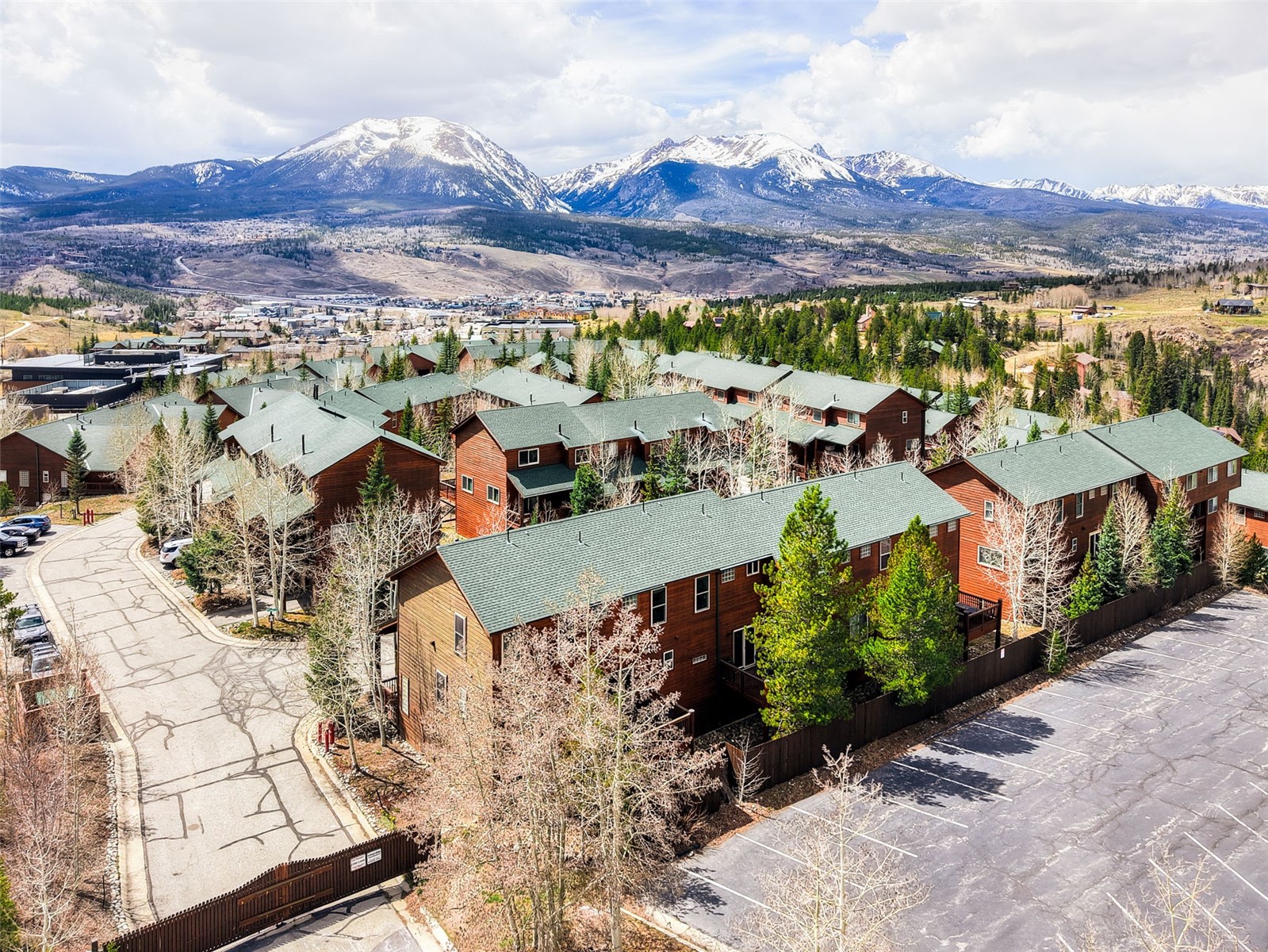 This high-angle aerial view captures a multi-unit residential complex featuring brown wood siding and dark green shingled roofs. The property is nestled in a mountainous landscape with snow-capped peaks in the background and a paved parking area in the foreground. The scene conveys a serene, alpine atmosphere, highlighting the integration of the buildings into the natural, wooded environment.