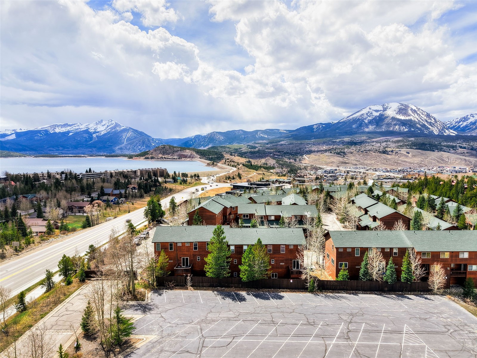 This high-angle aerial view captures a multi-unit residential complex featuring rustic wood siding and green shingled roofs, set against a dramatic backdrop of snow-capped mountains and a large lake. The foreground includes a paved parking lot, while a winding road leads past the property toward the distant town and mountain range. The scene conveys a serene, alpine atmosphere, highlighting the property's scenic location and proximity to outdoor recreational areas.