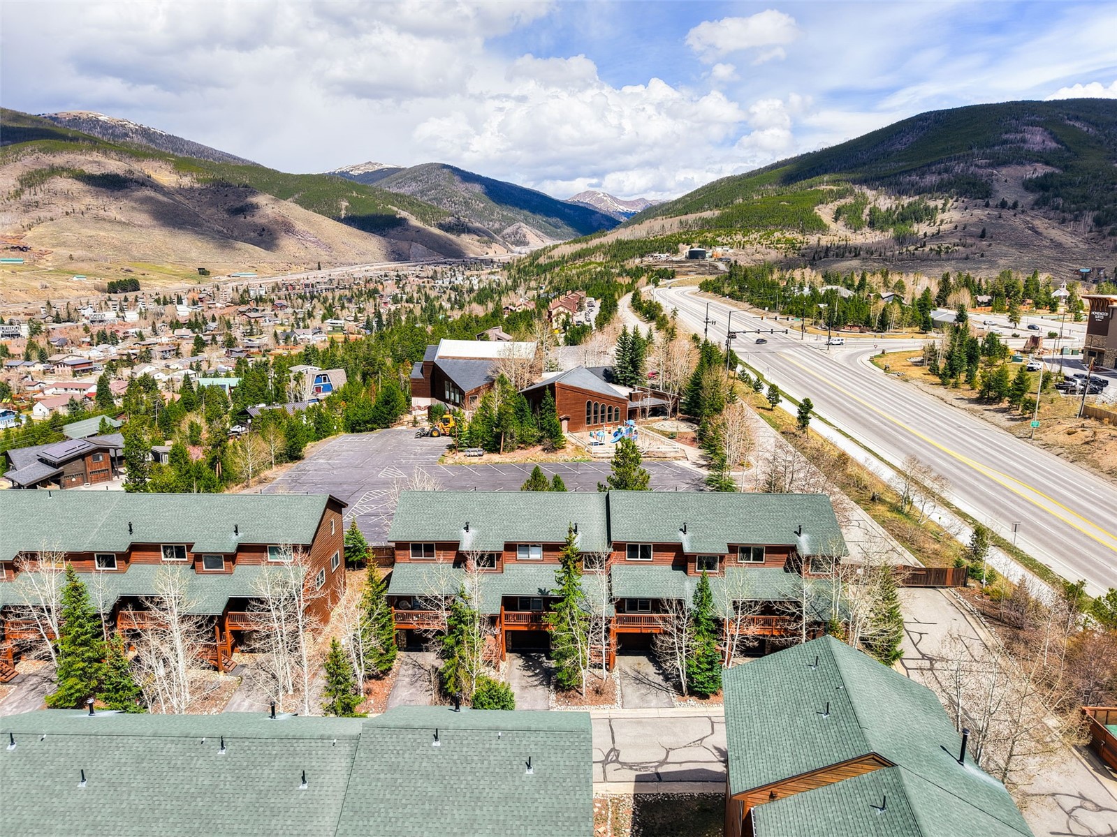 This high-angle aerial view captures a cluster of multi-story townhomes with dark green shingled roofs and wood-paneled siding, nestled in a mountainous landscape. The scene includes a paved parking area, a nearby commercial building, and a wide highway stretching into the distance toward rolling hills and a cloudy sky. The perspective provides a comprehensive look at the property's integration into the surrounding alpine environment.