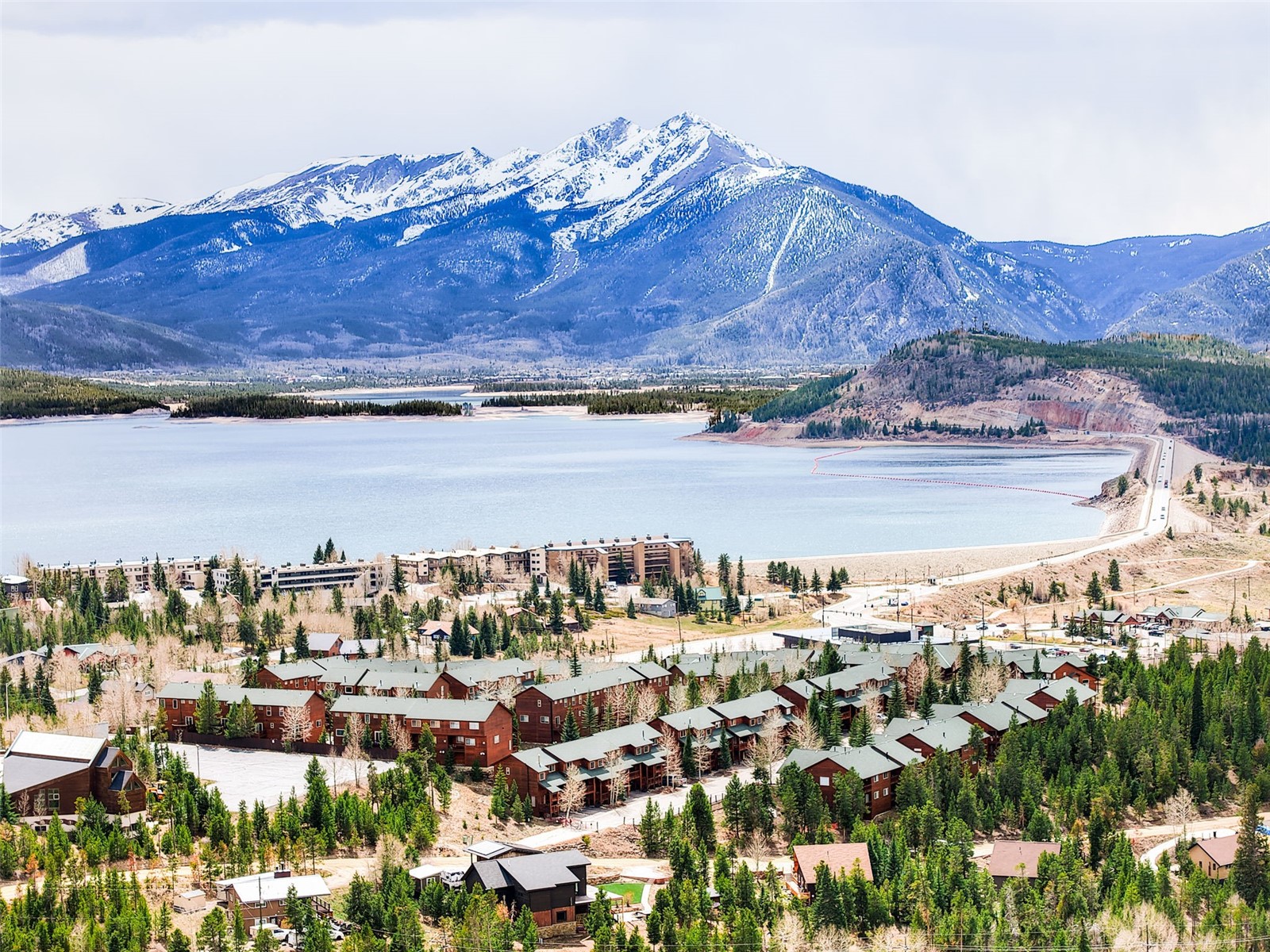 This high-angle aerial view captures a scenic mountain community nestled along the shores of a expansive, calm lake. The landscape features a dense forest of evergreen trees surrounding rows of multi-story residential townhomes with dark-toned roofs. In the background, a majestic, snow-capped mountain range dominates the horizon, creating a dramatic and serene backdrop for this lakeside development.