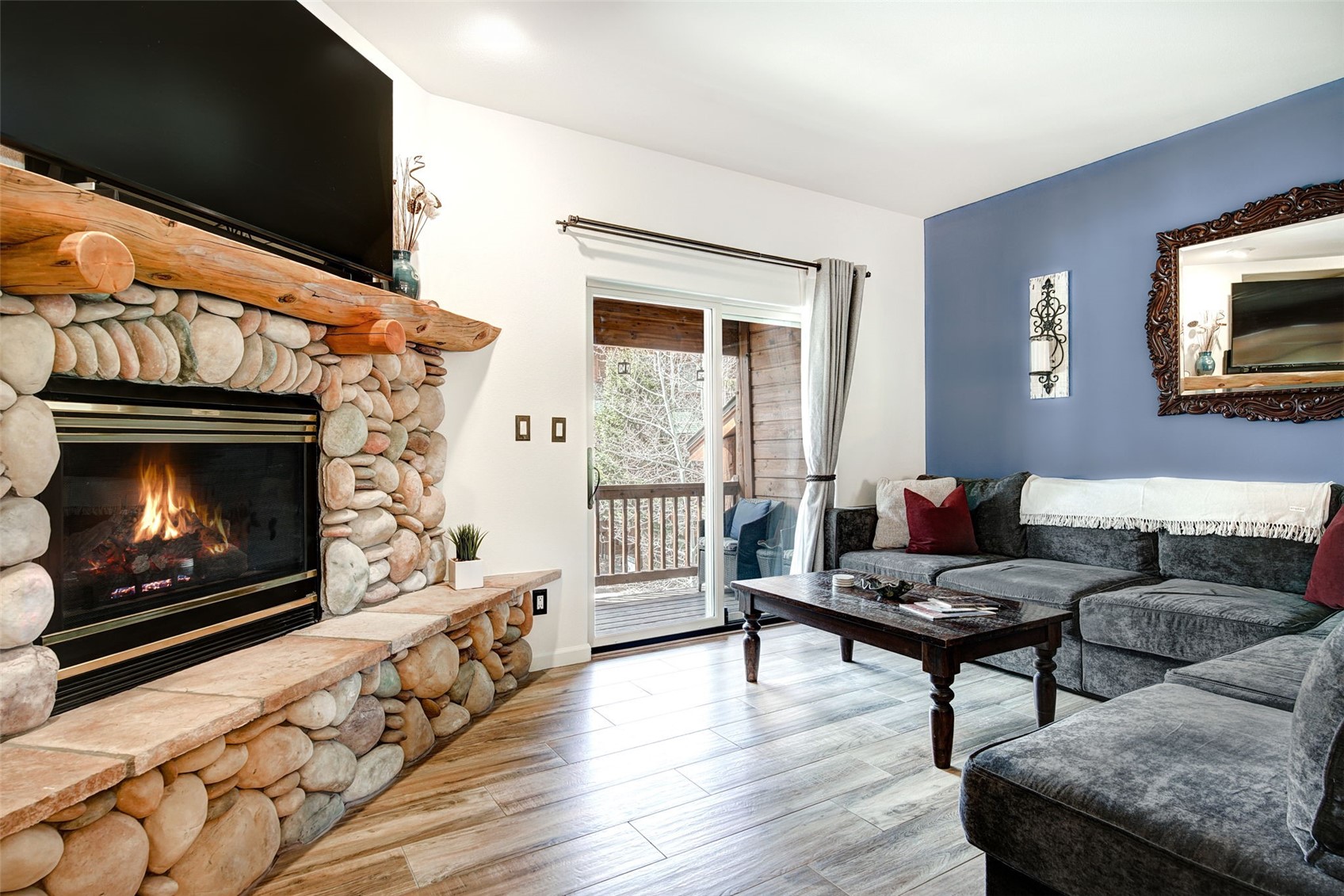 This cozy living room features a prominent stone-clad fireplace with a rustic timber mantel and a large television mounted above. A comfortable dark gray sectional sofa sits opposite the fireplace, complemented by a dark wood coffee table and a striking blue accent wall adorned with a decorative mirror. The space is brightened by natural light streaming through the sliding glass door that leads to an outdoor deck, all set against a backdrop of wood-look tile flooring.