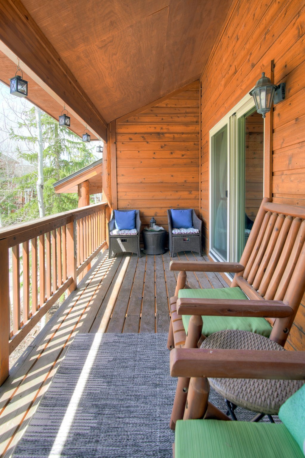 This elevated wooden balcony offers a cozy outdoor retreat with a rustic aesthetic, featuring warm-toned wood siding and a matching wooden railing. The space is furnished with a pair of wooden chairs with green cushions in the foreground and two wicker chairs with blue cushions positioned against the wall, separated by a small side table. The perspective captures the length of the deck, highlighting the natural light filtering through the trees and the inviting, relaxed atmosphere of the outdoor living area.