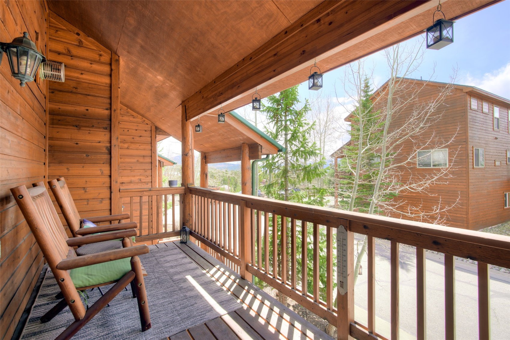 This covered wooden balcony features two rustic rocking chairs placed on a patterned rug, offering a cozy outdoor seating area. The space is framed by warm-toned wood siding and a wooden railing, with several hanging lanterns providing ambient lighting. From this elevated perspective, the deck overlooks a quiet residential street and surrounding trees, creating a peaceful and inviting atmosphere.