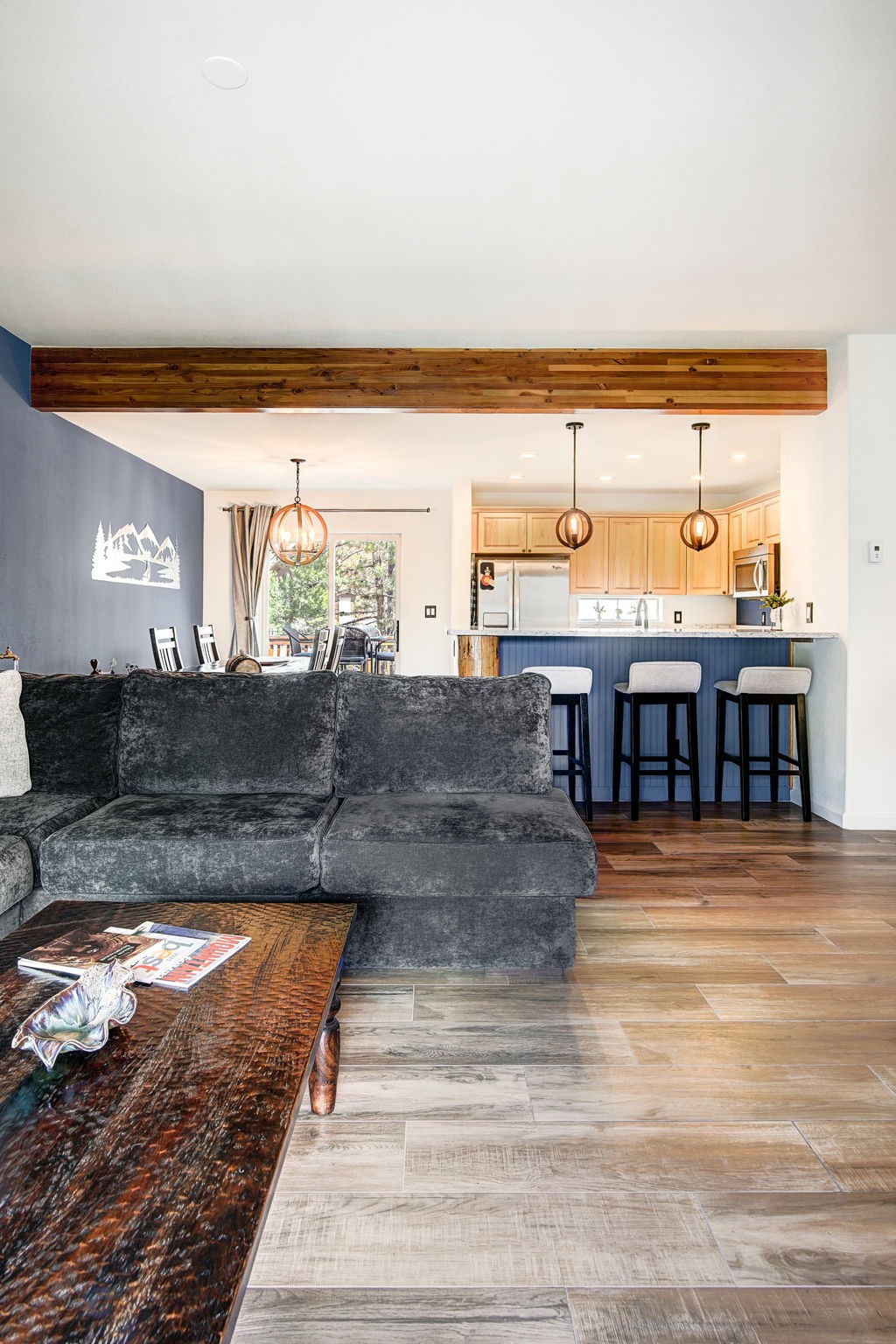 This open-concept living space features a large, dark-toned sectional sofa facing a rustic, textured coffee table. A prominent wood beam spans the ceiling, separating the living area from the kitchen, which is equipped with light wood cabinetry and a blue-painted island with bar seating. The room is finished with wood-look flooring and modern pendant lighting, creating a warm and inviting atmosphere.