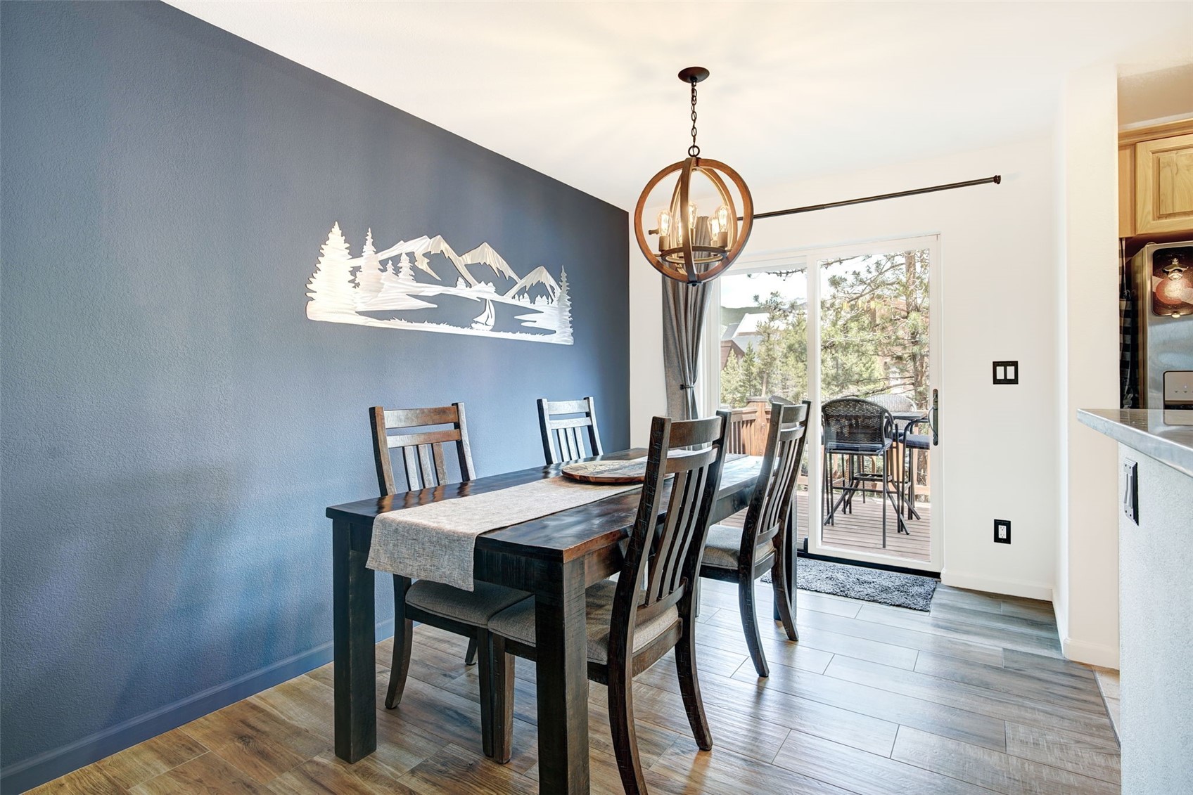 This dining area features a dark wood table set with a neutral runner, surrounded by four matching chairs. A striking metal mountain-themed wall art piece serves as a focal point on the deep blue accent wall, while a rustic orb-style chandelier hangs above. A sliding glass door provides natural light and a view of the outdoor deck, creating an inviting and cohesive space.