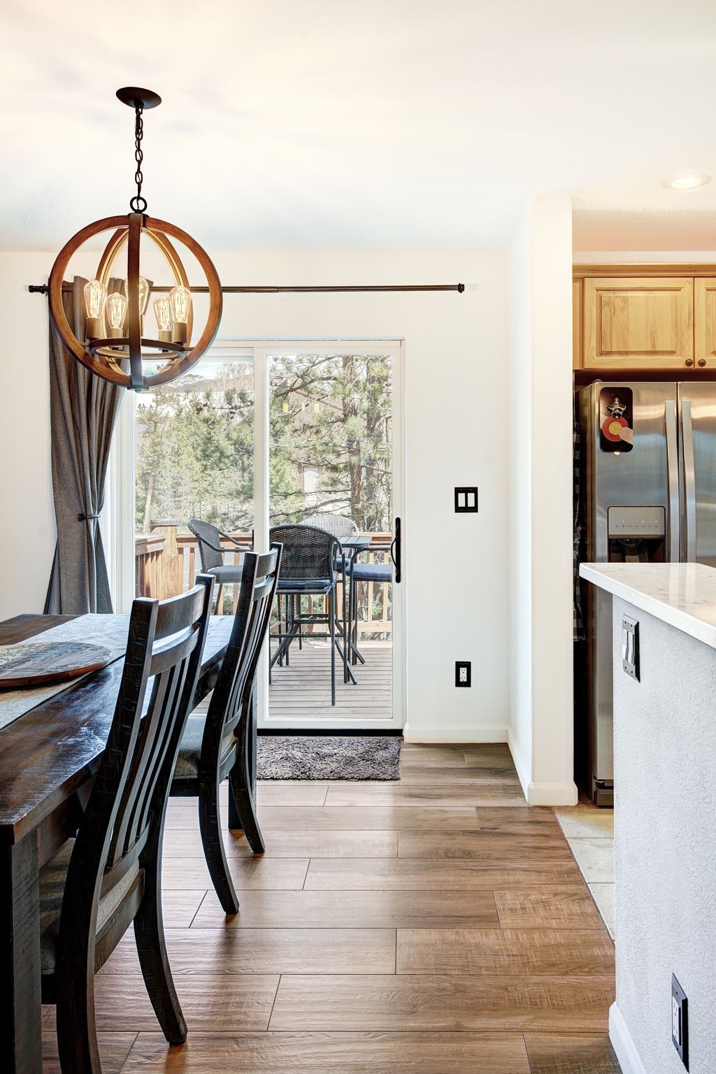 This dining area features a dark wood table paired with matching chairs, illuminated by a prominent spherical wooden chandelier. The space flows seamlessly into a kitchen area with stainless steel appliances and light wood cabinetry, while a sliding glass door provides a view of an outdoor deck. The room is finished with warm-toned wood-look flooring, creating a cohesive and inviting atmosphere.