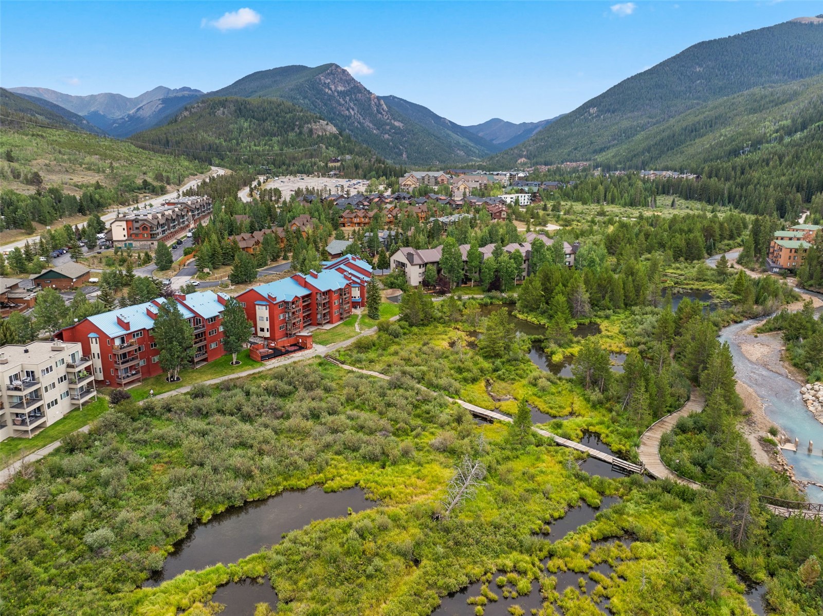 This high-angle aerial view captures a scenic mountain resort community nestled in a lush valley. The scene features multi-story residential buildings with distinctive blue roofs, a winding wooden boardwalk traversing a vibrant wetland area, and a clear stream flowing through the landscape. The backdrop is dominated by majestic, tree-covered mountains under a bright, clear sky, emphasizing the tranquil and natural setting of the property.