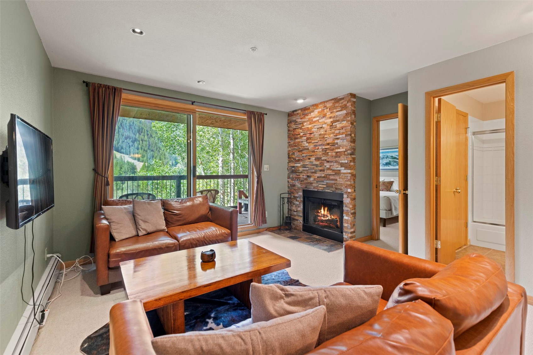 This cozy living room features a warm, rustic aesthetic with two brown leather sofas arranged around a wooden coffee table. A prominent stone-clad chimney breast serves as the focal point, housing a glowing fireplace that adds to the inviting atmosphere. Large sliding glass doors provide a view of the lush green trees outside, while a doorway leads to an adjacent bedroom and bathroom area.