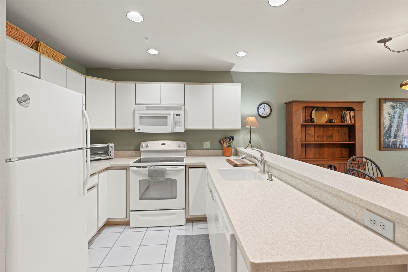 This kitchen features a functional U-shaped layout with white cabinetry, a white appliance suite, and light-colored countertops. A breakfast bar extends from the counter, providing a transition into the adjacent dining area where a wooden hutch is visible. The space is illuminated by recessed ceiling lights and a small table lamp, creating a warm and inviting atmosphere.