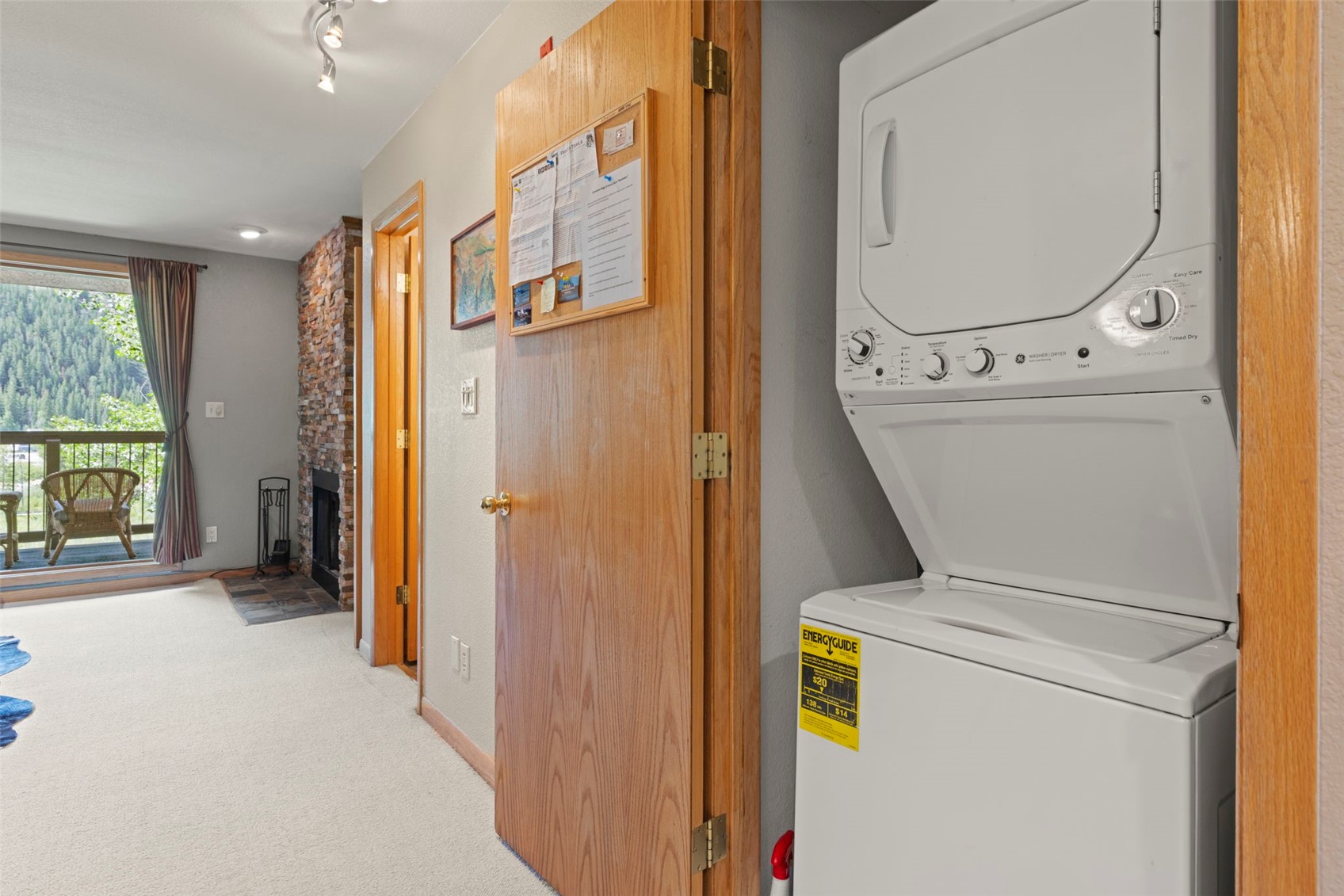 This interior view captures a laundry area featuring a stacked washer and dryer unit positioned next to a wooden door with a cork bulletin board. To the left, a hallway leads toward a living space with a stone-clad chimney breast and a sliding glass door that opens onto a balcony with outdoor seating. The neutral color palette and carpeted flooring create a cohesive transition between the utility space and the main living area.