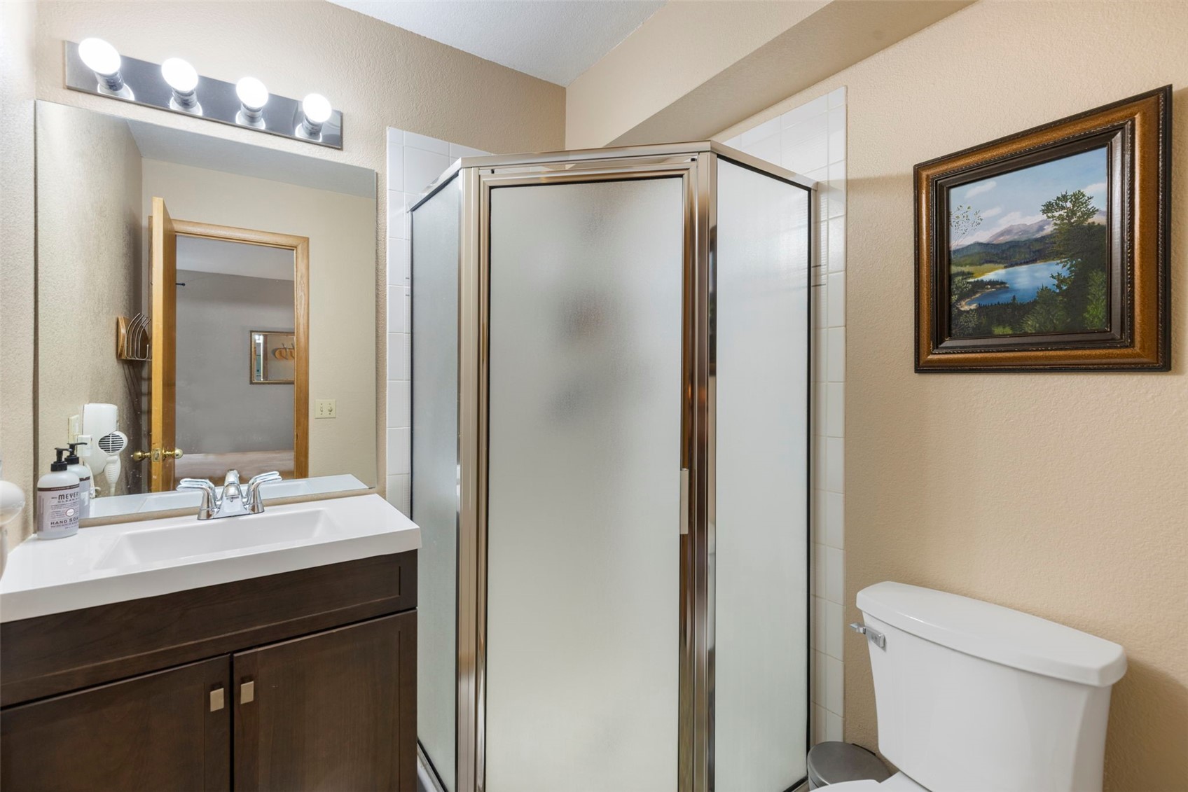 This compact bathroom features a dark wood vanity with a white sink and a chrome faucet, positioned next to a corner shower stall with frosted glass panels. A large mirror above the vanity reflects the room, while a framed landscape painting hangs on the adjacent wall near the white toilet. The space is illuminated by a four-bulb vanity light fixture, creating a functional and tidy atmosphere.