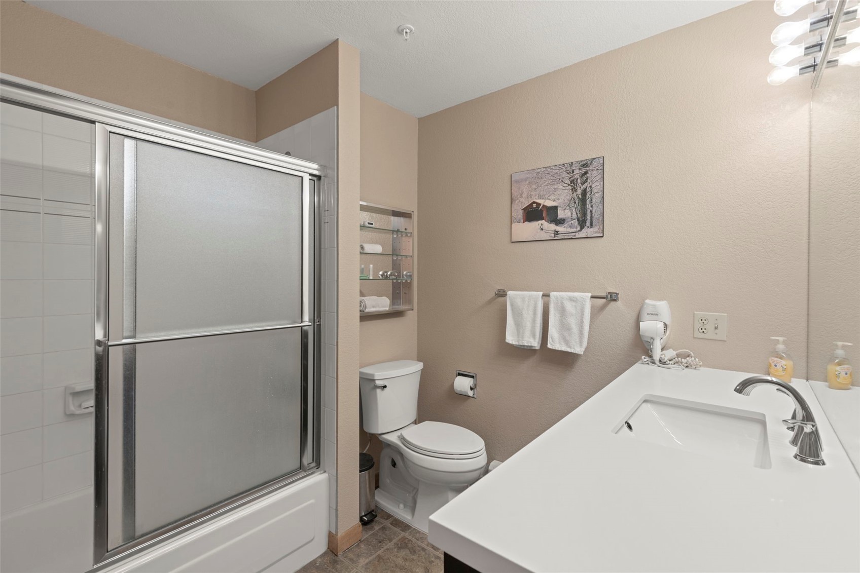 This functional bathroom features a white vanity with a rectangular sink, a shower-tub combination with a sliding glass door, and a standard toilet. The walls are painted a neutral beige, complemented by a small framed landscape print and a wall-mounted storage rack. The space is illuminated by a multi-bulb vanity light fixture, creating a clean and practical layout.