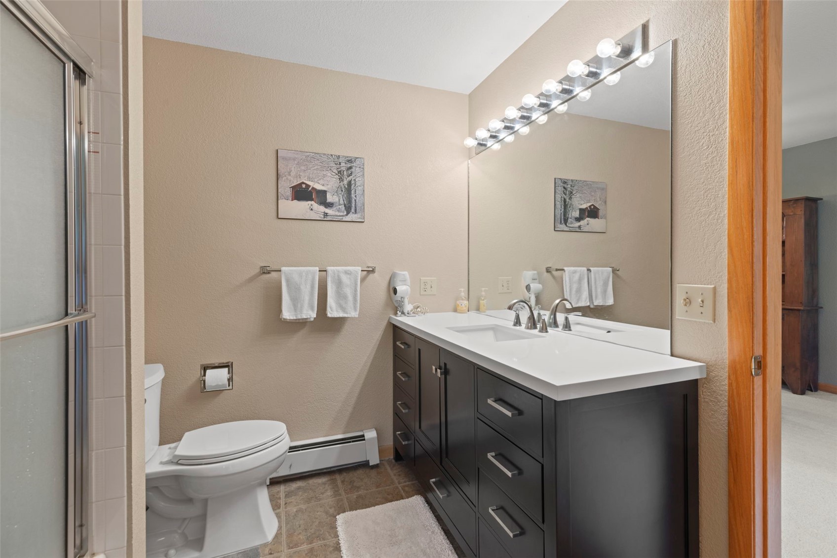This bathroom features a dark-toned vanity with a white countertop and a large, full-width mirror illuminated by a multi-bulb vanity light. To the left, a white toilet sits beside a shower enclosure with a glass door, while a small rug rests on the tiled floor. The walls are painted a neutral tan, accented by a framed winter landscape print above the towel rack.