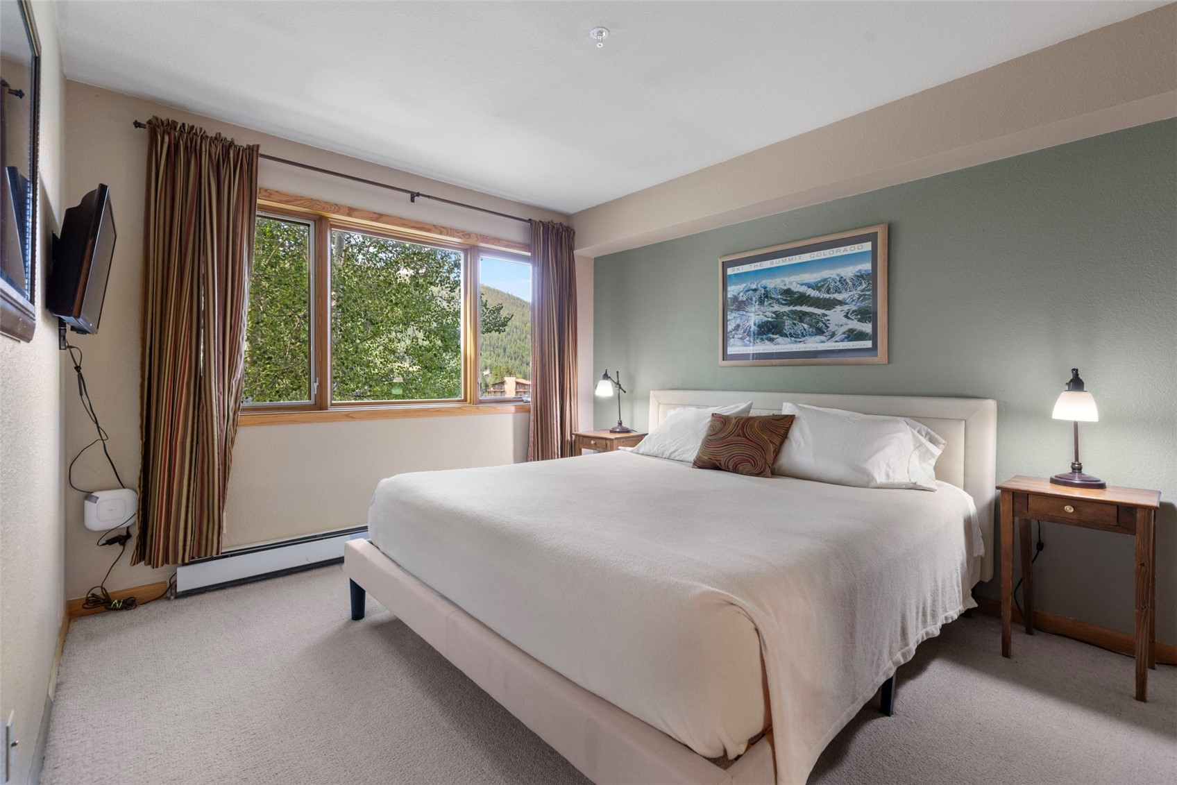 This bedroom features a neutral color palette with a soft green accent wall behind a bed dressed in white linens. A large window with striped curtains allows natural light to fill the room, offering a view of the surrounding trees and mountains. The space is completed by two wooden nightstands with matching lamps and a framed mountain landscape print above the headboard.