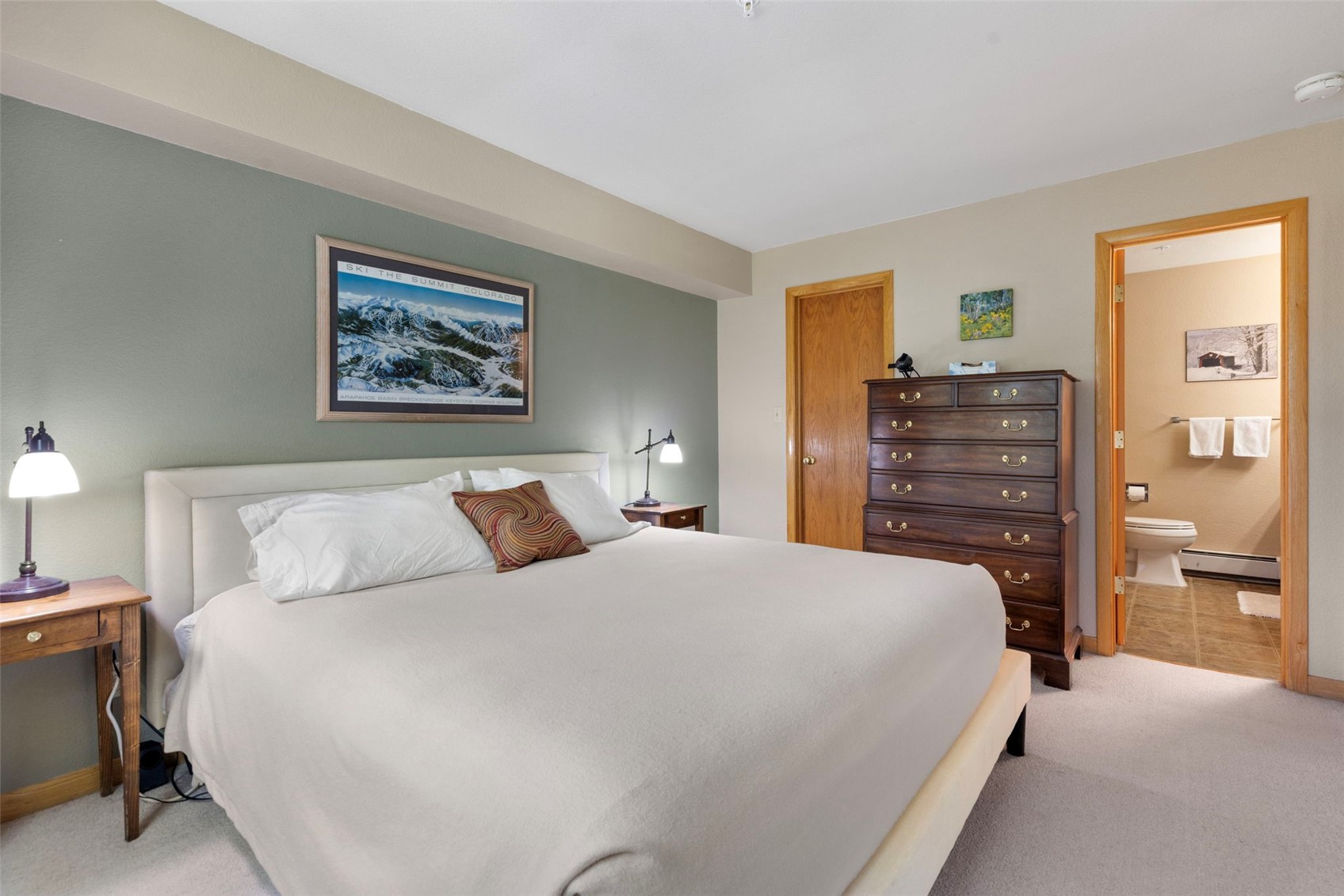 This bedroom features a neutral-toned bed with a light-colored upholstered headboard, centered against a sage green accent wall adorned with a framed mountain landscape print. To the right, a tall, dark wood dresser provides storage, while a doorway leads into an attached bathroom with a visible toilet and towel rack. The room is illuminated by two bedside lamps, creating a cozy and inviting atmosphere.