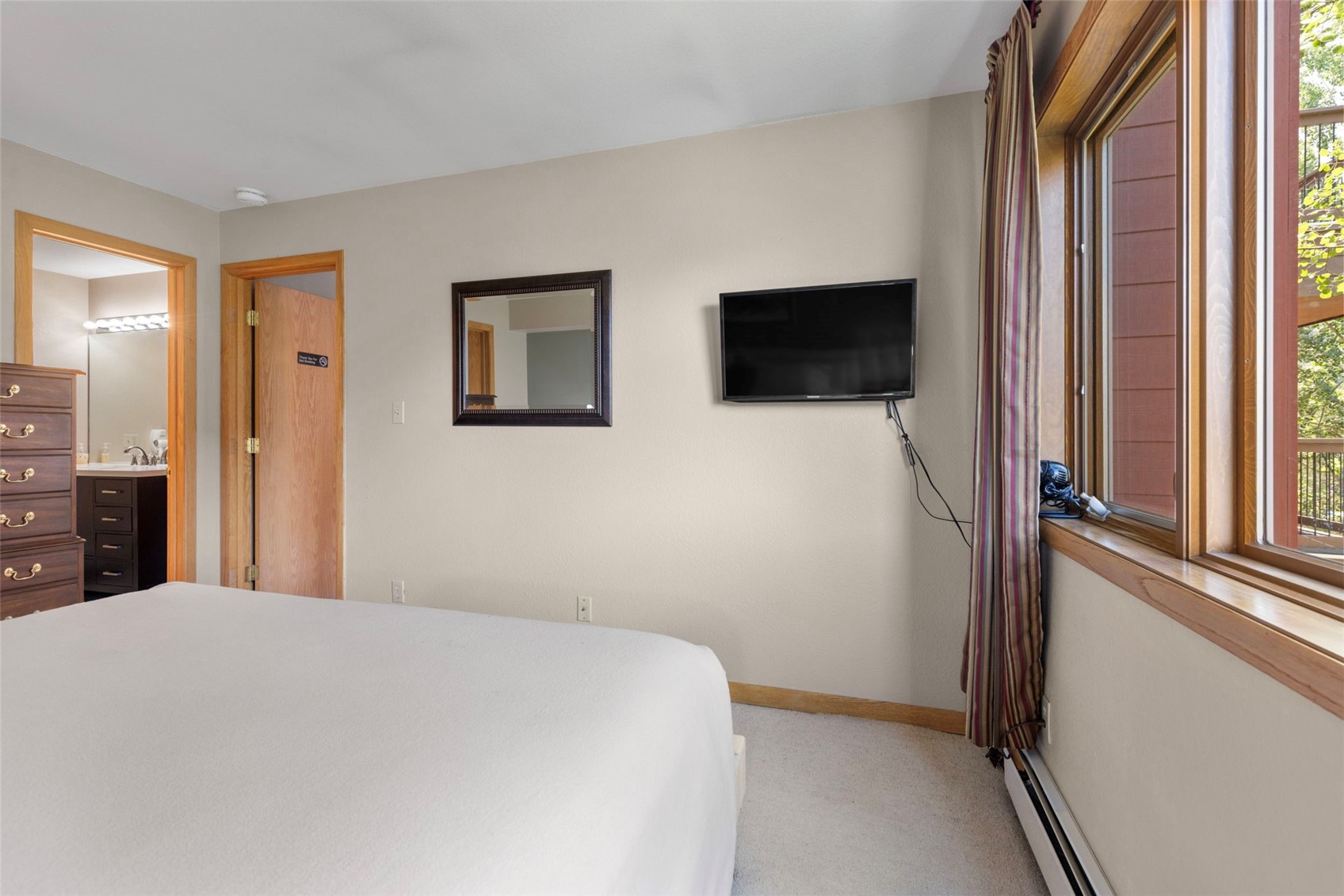 This bedroom features a neutral color palette with light-toned walls and carpeted flooring. The room includes a wall-mounted television, a framed mirror, and a wooden dresser, with an open doorway leading into an adjacent bathroom. A window with striped curtains provides natural light and a view of an exterior balcony.