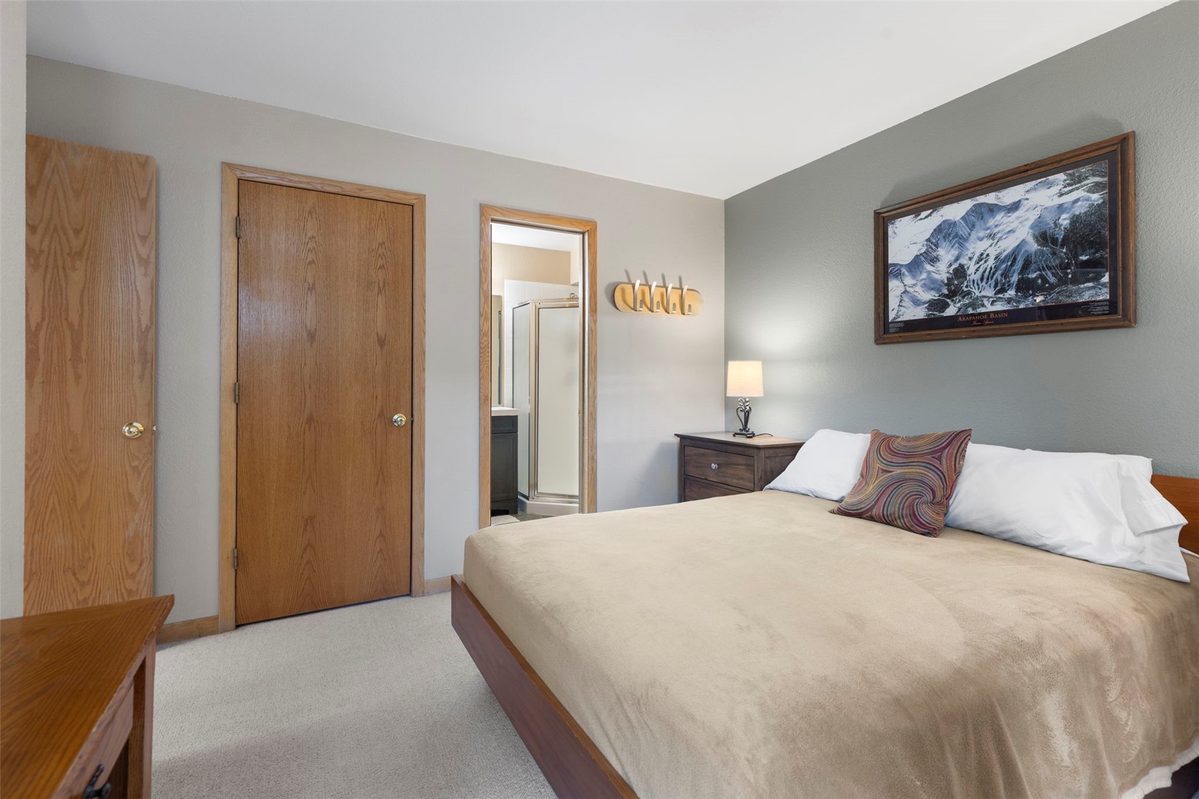This bedroom features a neutral color palette with a light-colored bedspread and a patterned accent pillow. A wooden nightstand with a lamp sits beside the bed, while a framed mountain landscape print hangs on the wall above. The room includes a doorway leading to an adjacent bathroom and a wooden door leading to a closet, creating a functional and cozy living space.
