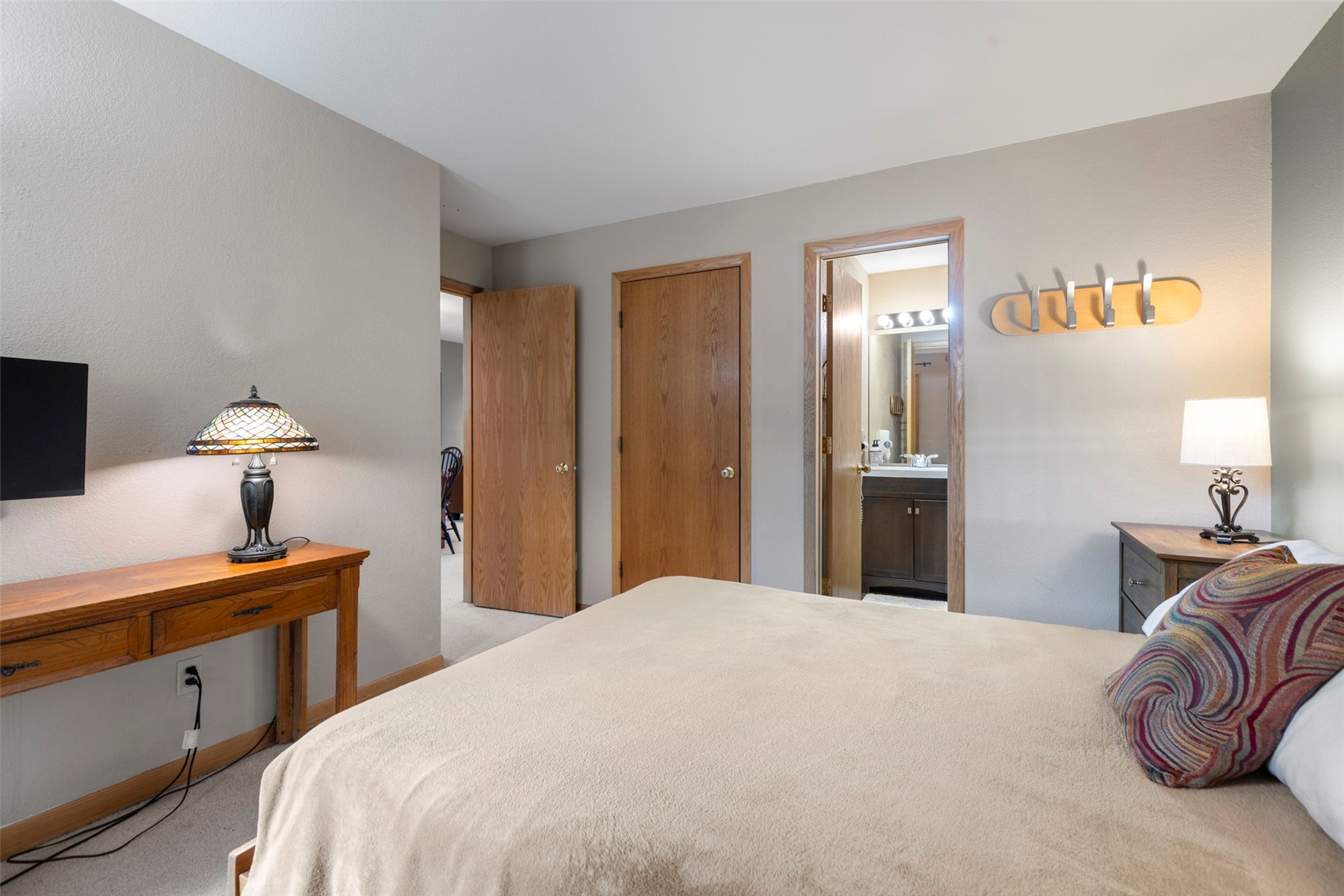 This bedroom features a neutral color palette with a tan bedspread and a patterned accent pillow. The room includes a wooden desk with a stained-glass style lamp, a wall-mounted television, and a wooden coat rack on the wall. A doorway leads into an adjacent bathroom, while another door provides access to a hallway, creating a functional and well-connected layout.