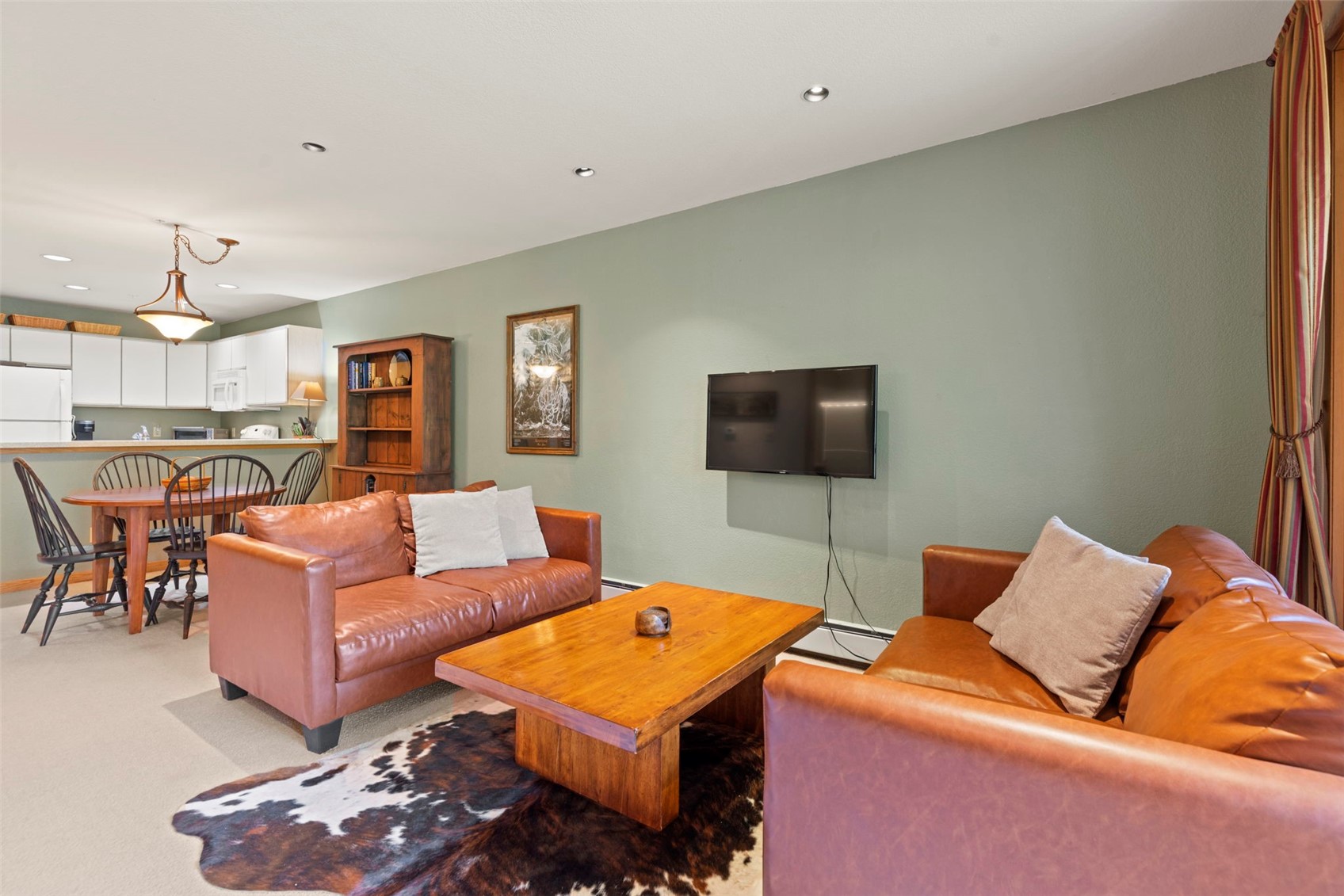 This open-concept living area features a warm, rustic aesthetic with two brown leather sofas arranged around a solid wood coffee table atop a cowhide rug. The space is painted in a soft sage green, with a wall-mounted television and a tall wooden bookshelf providing functional storage. In the background, a dining area with a wooden table and chairs transitions into a kitchen, creating a cohesive and inviting living space.